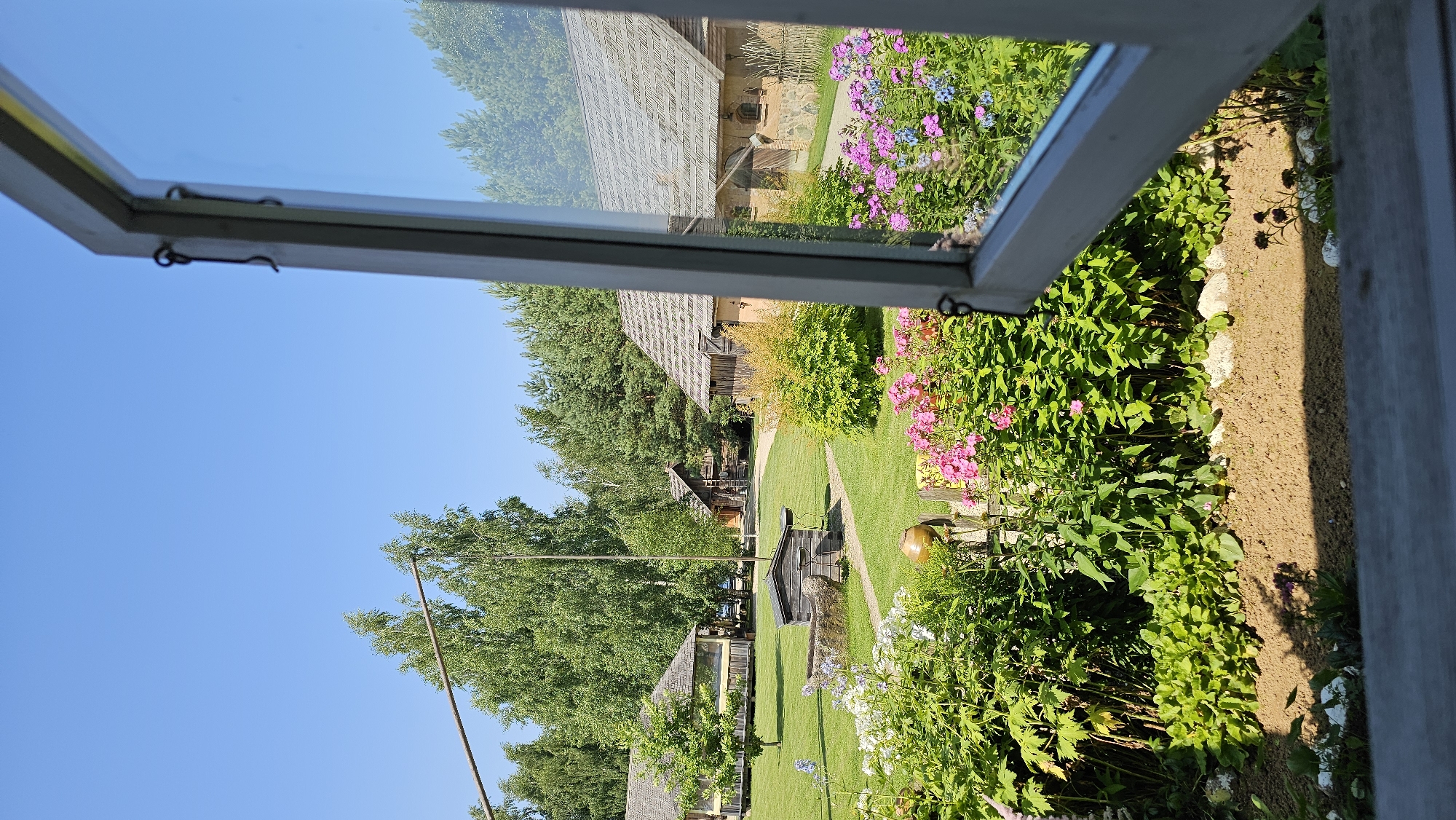 A photograph taken from inside a building looking out through an open window frames a pastoral scene in Sodeliškiai, Lithuania. The window, with its white-painted wooden frame and metal latch, is partially open and angled to the right. The foreground of the image is dominated by a lush garden bed filled with vibrant pink and blue flowers, including phlox, and various green leafy plants. Beyond the garden, a manicured green lawn stretches towards several rustic wooden buildings with thatched roofs. In the middle ground, a traditional well with a stone base and a wooden structure sits on the lawn, accompanied by a tall wooden pole. The background features a dense forest of green trees under a clear blue sky. The overall impression is one of a tranquil rural setting.