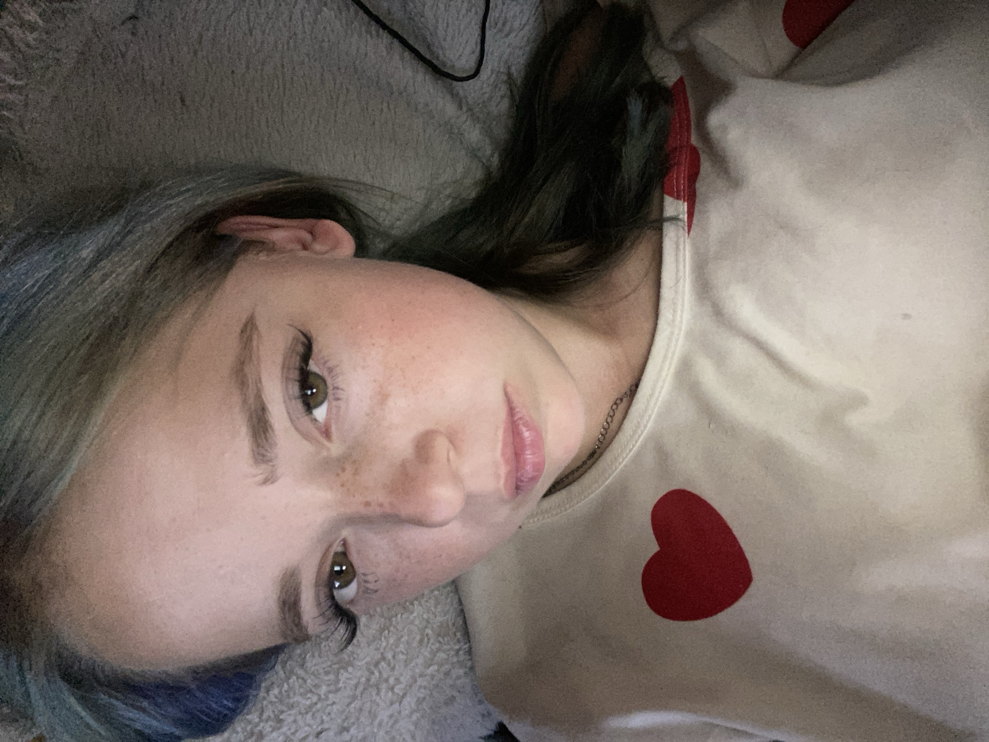The image is a close-up, low-angle, portrait-oriented photo of a young person lying down. They have light skin with freckles scattered across their cheeks. Their eyes are a hazel-brown color, and they are wearing dramatic false eyelashes. Their eyebrows are lightly filled in. The person has light brown hair with subtle blue and green highlights, and it appears to be styled down. They are wearing a light-colored, possibly cream or beige, long-sleeved shirt adorned with multiple red heart designs. The fabric of the shirt appears to be soft and possibly textured, with visible folds and creases. A thin silver chain necklace is visible around their neck. The background is a soft, textured surface, likely a blanket or bedding, in a muted grey tone. The lighting is dim, suggesting it might be nighttime or an indoor setting with low light. There is a faint dark cord visible in the upper left portion of the background. The overall emotion conveyed is relaxed and possibly contemplative. There is no visible text in the image. The provided location context is Nova Praha, Ukraine, but there are no visual cues in the image to confirm this location.