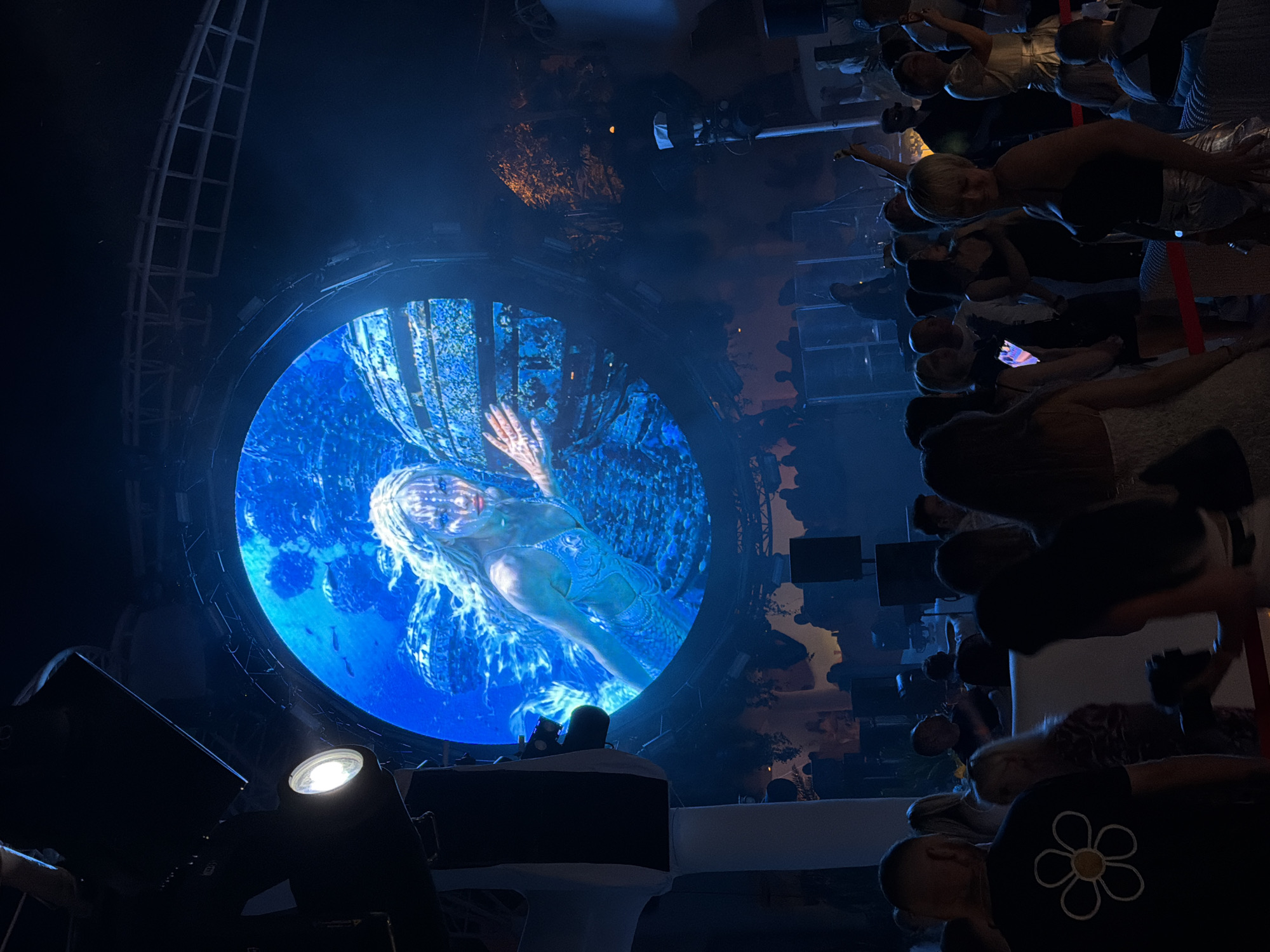 The image depicts a nightclub or event venue in Odesa, Ukraine, with a large circular LED screen as the central focal point. The screen displays a luminous image of a woman with blonde hair, adorned with body paint resembling tribal markings, in what appears to be an underwater or fantasy setting. She is wearing a light-colored, ornate costume. The screen is surrounded by stage lighting and rigging. In the foreground and midground, a diverse crowd of people are visible, some looking towards the screen, others engaged in conversation or dancing. They are dressed in casual and club attire. The environment is dimly lit, with blue and white lighting illuminating the scene, creating a festive and energetic atmosphere. The overall impression is that of an entertainment event, likely a party or performance, held in a modern venue within Odesa.