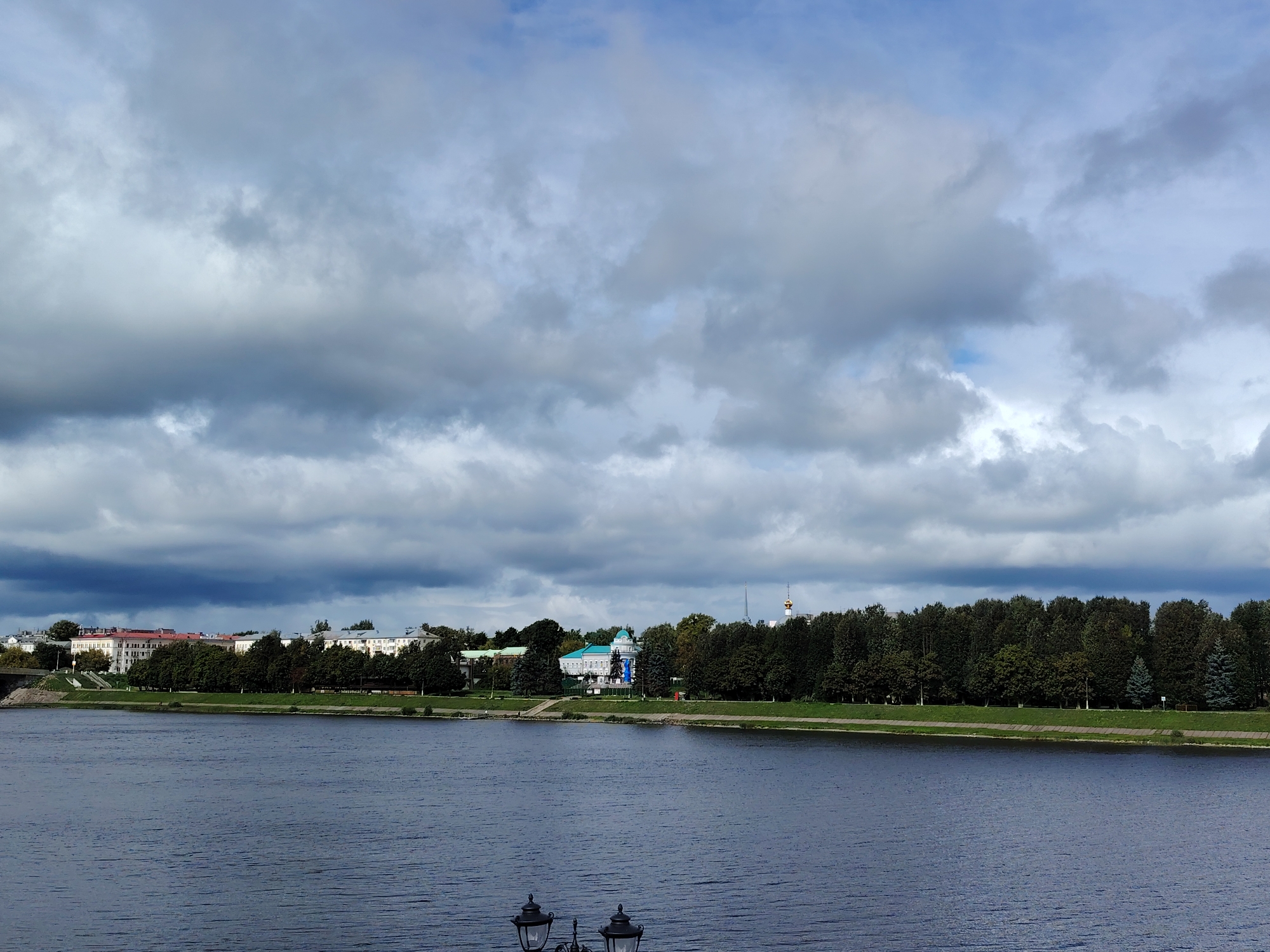 The image depicts a view across a wide river towards a city skyline under a cloudy sky. The river dominates the foreground, with its dark, rippling surface reflecting the overcast heavens. Along the far bank, a line of trees forms a green buffer between the water and the urban landscape. Behind the trees, buildings of varying architectural styles and heights are visible. Some buildings are light-colored and appear to be multi-story, while others are darker. In the middle ground, a prominent white building with a turquoise roof stands out, possibly a historical or public building. The sky is filled with dramatic, dark gray clouds, suggesting an impending storm or overcast weather. In the extreme foreground, the tops of two ornate black streetlights are visible, suggesting this is a public promenade or viewing area. The location is identified as the city of Tver, Russia, looking across the Volga River. No people are distinctly visible in the scene.