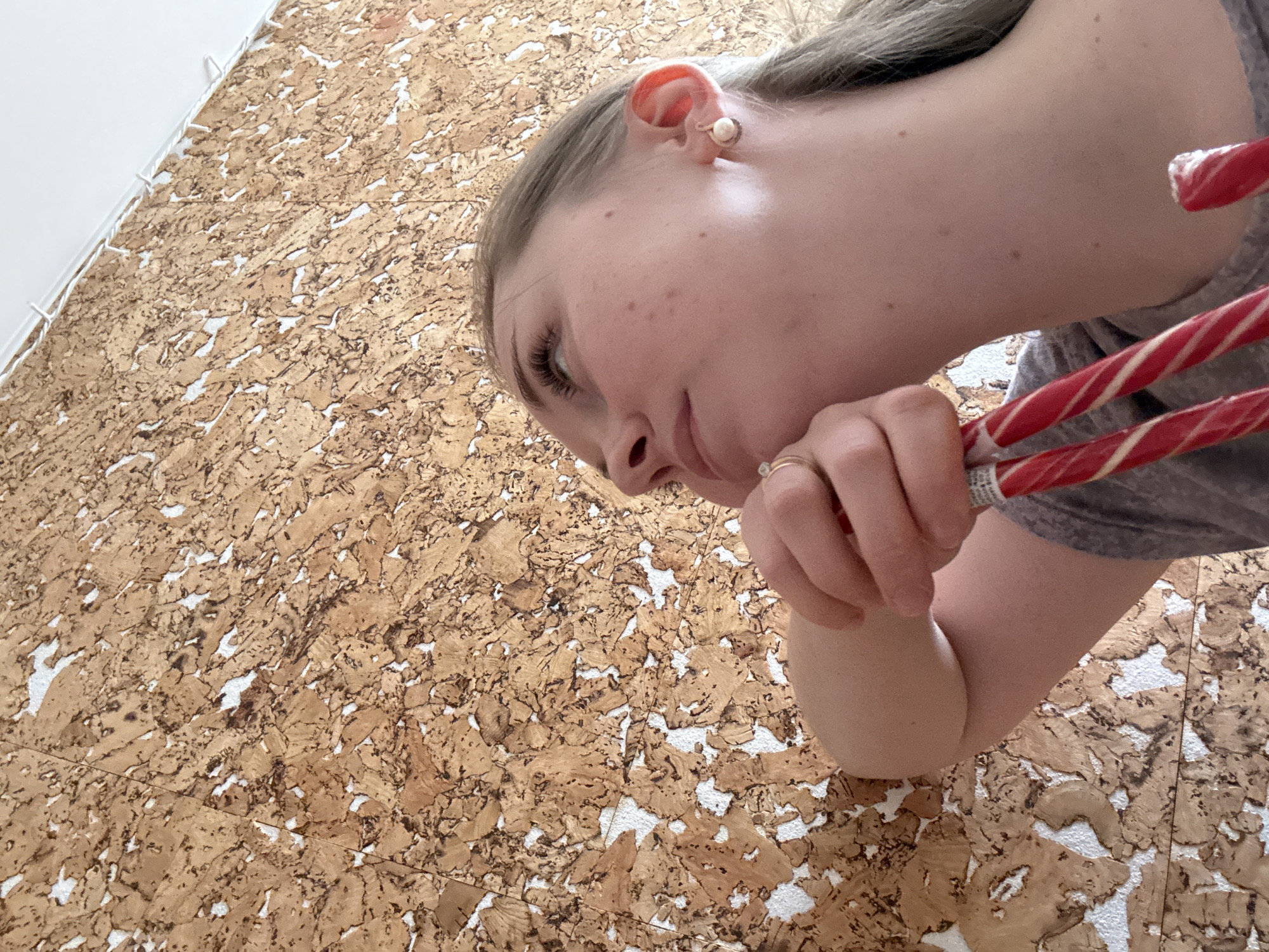 The image presents a close-up, diagonal view of a young person, likely female, from the upper torso upwards. She has fair skin with scattered freckles on her cheek and forehead, and light-colored hair that appears to be light brown or blonde, pulled back. A single pearl stud earring is visible in her right earlobe. On the ring finger of her right hand, she wears two rings: a simple gold band and another with a small, light-colored stone. She is wearing a casual gray short-sleeved top.
Her right arm is bent, with her elbow resting on a textured surface, and her hand holds two red and white striped candy canes or similar lollipops. The person is looking downwards and slightly to the left, with a neutral to mildly pensive expression, suggesting focus or contemplation on something out of frame below.
The background is dominated by a brown and white textured surface resembling cork board or flooring. It's composed of numerous irregularly shaped pieces of light brown cork with distinct white spaces or gaps between them. In the upper left portion of the image, a solid white surface, possibly a wall or a large bound object, meets the cork. This transition is marked by a line of small, white, cord-like elements, possibly binding or decorative trim. The lighting is soft and even, indicating an indoor setting. Small, illegible text is faintly visible on the wrappers of the candy canes.