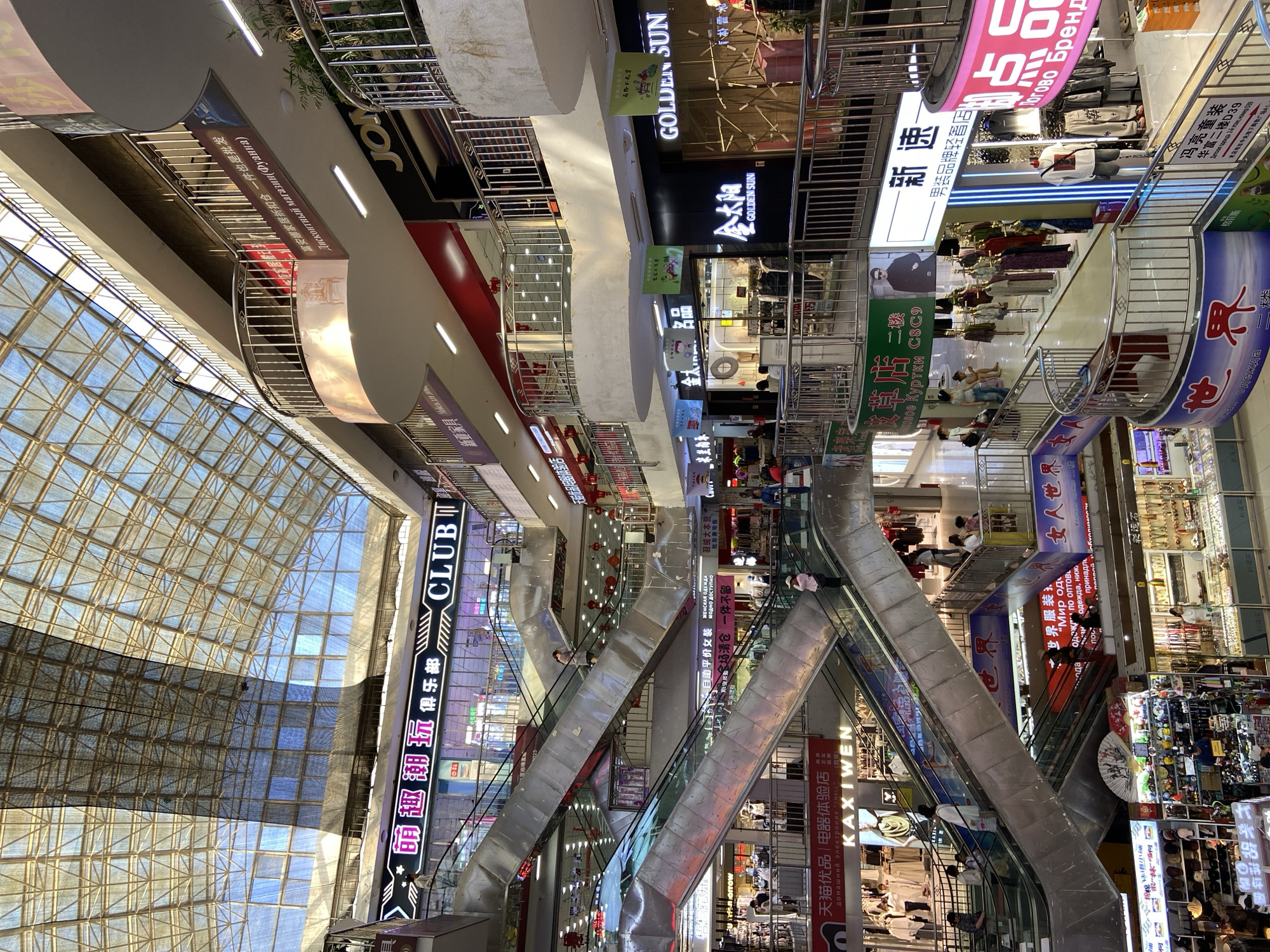 The image shows the interior of a multi-level shopping mall in Xing'an Subdistrict, China. The mall features escalators and walkways connecting various floors. Several shops are visible, displaying merchandise such as clothing, electronics, and accessories. Signage in Chinese and Russian indicates the presence of diverse retail outlets. People are seen walking on escalators, browsing in shops, and interacting within the mall environment. The upper part of the mall is covered by a glass roof structure, allowing natural light to filter in. Red lanterns are hung in some areas, suggesting festive decorations.