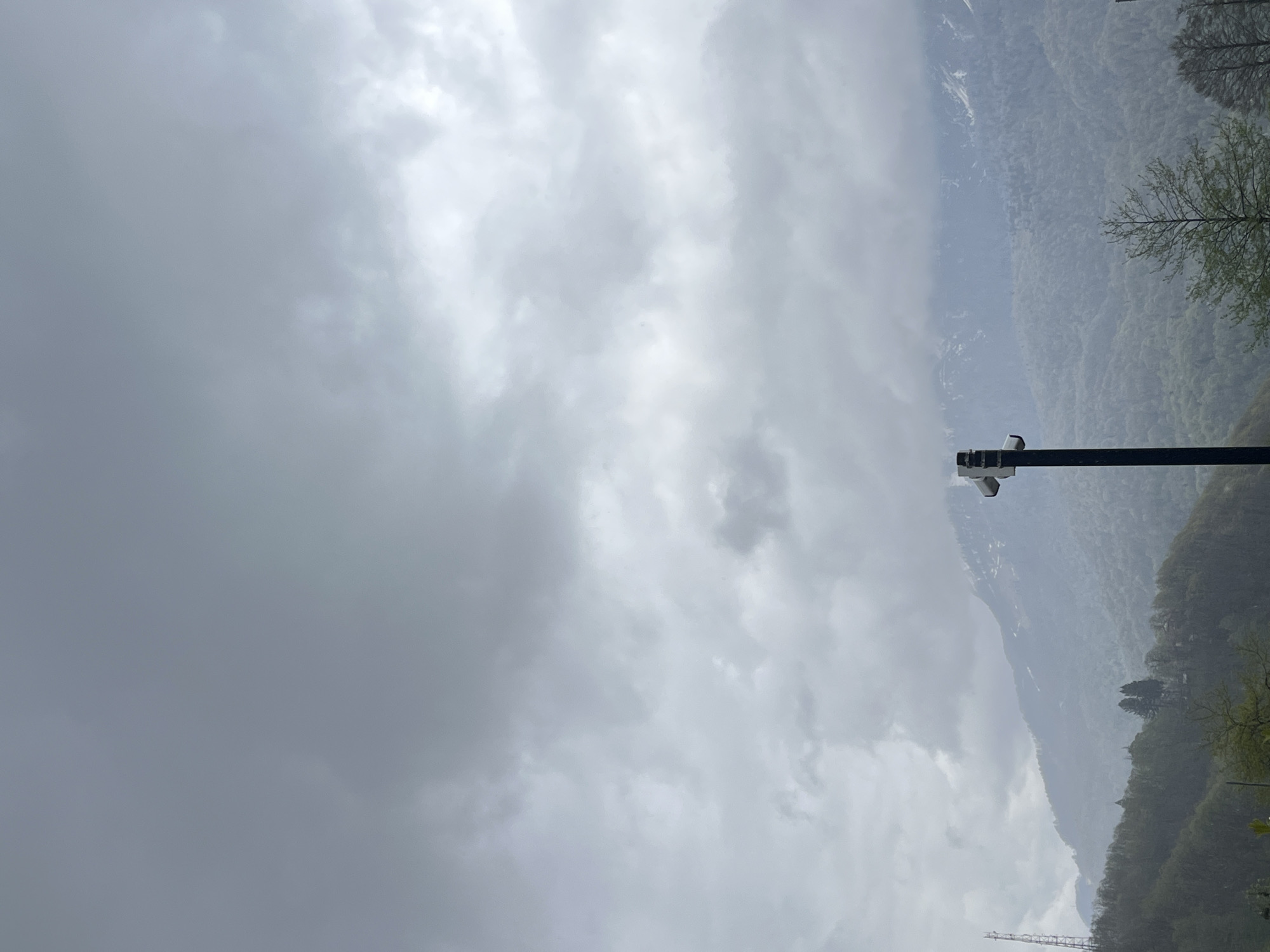 The scene is a cloudy day in a mountainous area. A black lamppost is on the right side of the image. In the background, there are forested mountains with visible cloud cover. The sky is predominantly gray. A cell tower can be seen towards the bottom of the image.