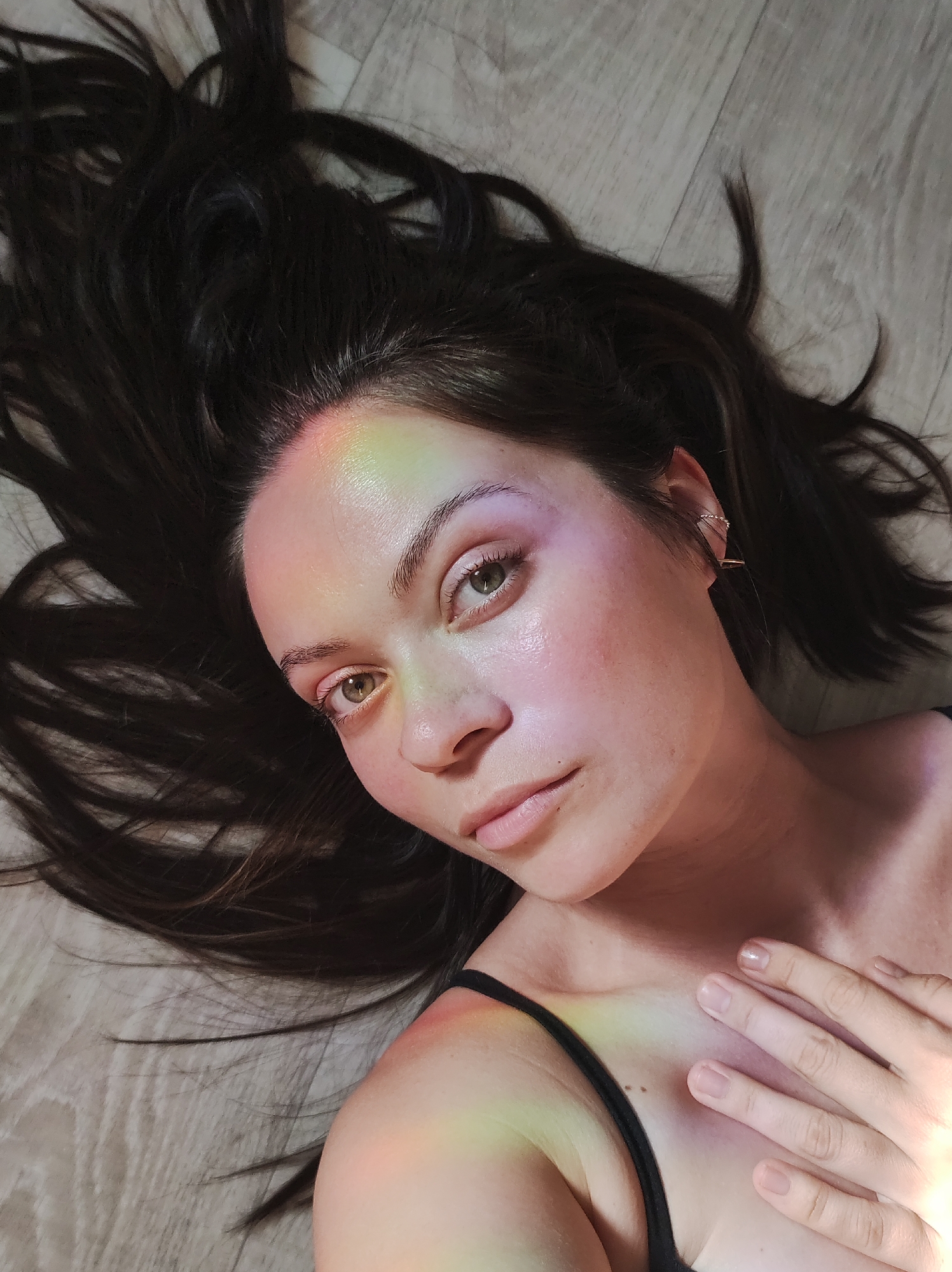 A young woman with fair skin and dark brown hair is lying on a light-colored, wood-look floor. Her hair is spread out around her head. She has light green eyes and is wearing a small earring on her left ear. Her left hand is resting on her chest. Rainbow light is reflecting across her forehead, face, chest and hand. She is wearing a black tank top with thin straps. The location is Unknown, Unknown.