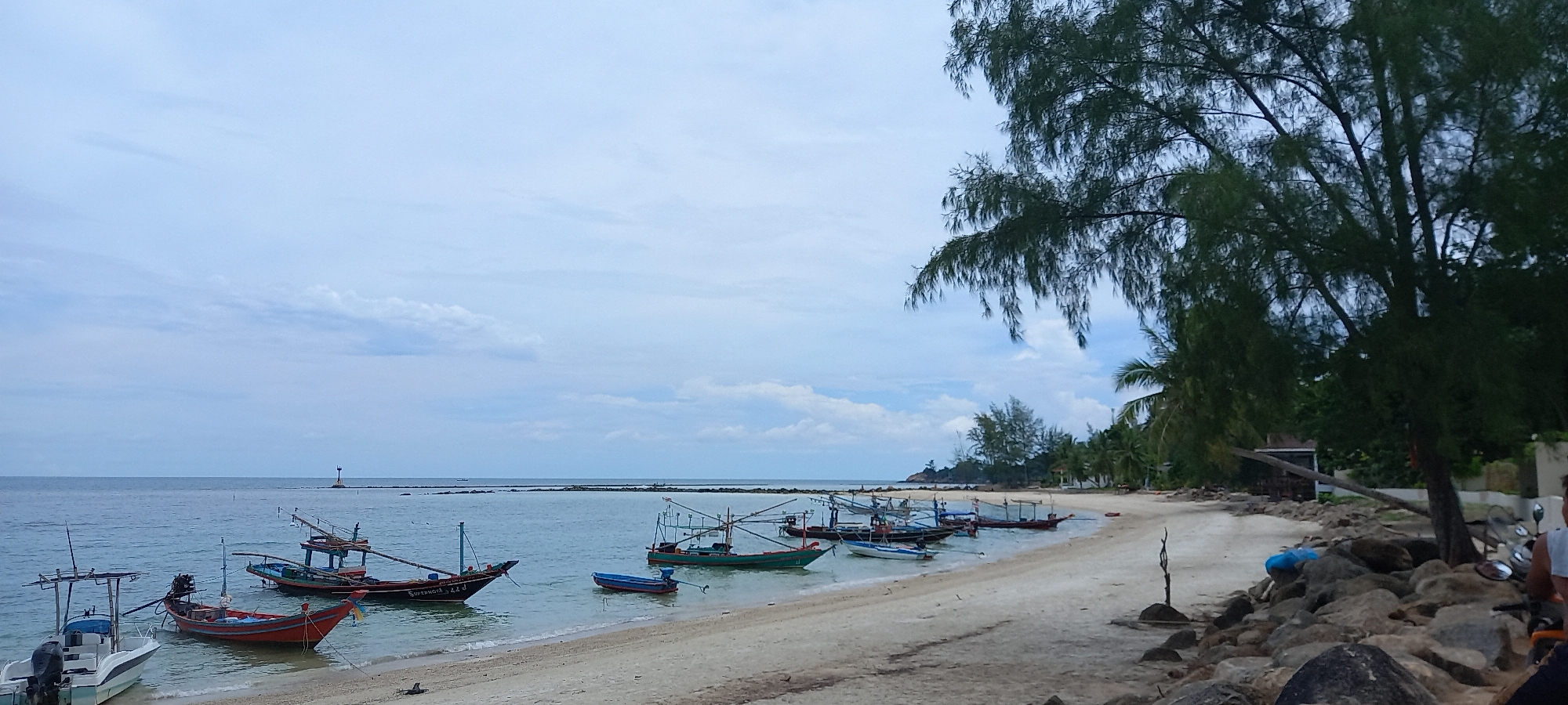 The image depicts a beach scene in Baan Chaloklum, Thailand. Several small fishing boats are moored along the shoreline, their prows facing the sea. The beach is composed of light-colored sand. A line of trees and vegetation borders the beach, transitioning into a rocky area in the foreground right. In the distance, a small structure, possibly a lighthouse, is visible on the horizon. The sky is overcast with light clouds.