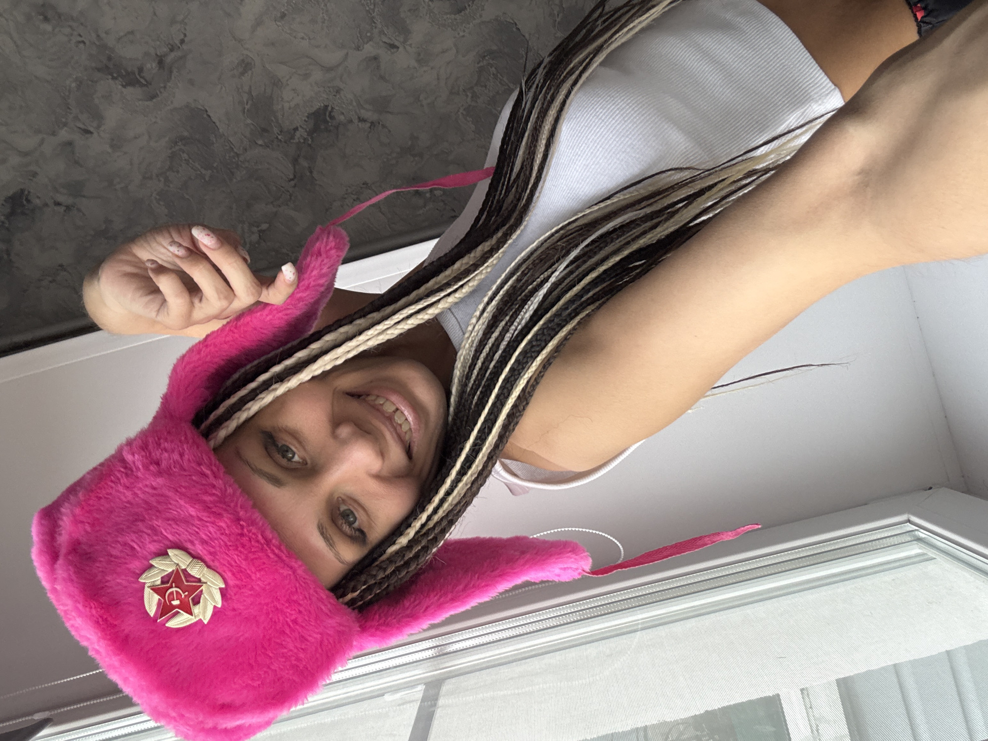 A young woman is photographed from below, wearing a white tank top and a bright pink ushanka hat emblazoned with a Soviet military star. She smiles slightly, her braided hair cascading around the hat and the top of the frame. The background consists of a blurred grey ceiling and a window with white frames. The location appears to be indoors.