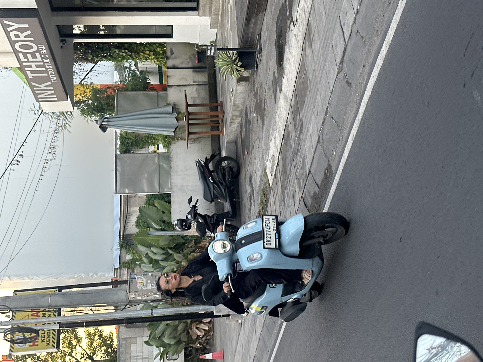 A woman is riding a light blue scooter down a paved street in North Kuta, Indonesia. She is wearing a black jacket, black pants, and sandals, with sunglasses on. Her hair is dark and long, and she is looking to her right. The scooter has a license plate that reads "DK2774FCW" and "07-28". In the background, there is a building with a sign that says "INK THEORY TATTOO STUDIO BALI". There is also a parked black scooter and an umbrella. The scene appears to be in an urban or commercial area.