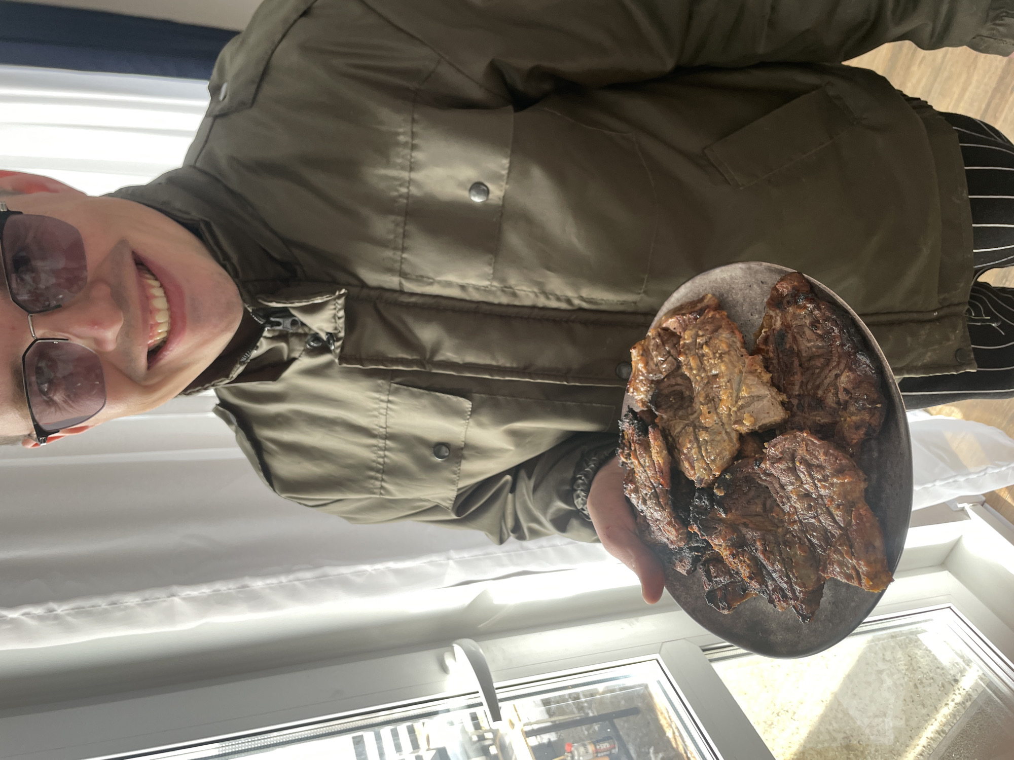 A man wearing a green jacket and sunglasses is holding a plate of cooked meat near a window. The window appears to be in a residential setting, possibly in Подшивалово, Russia, although this is not definitively verifiable from the image. The man is smiling and presenting the food. The image is slightly rotated.