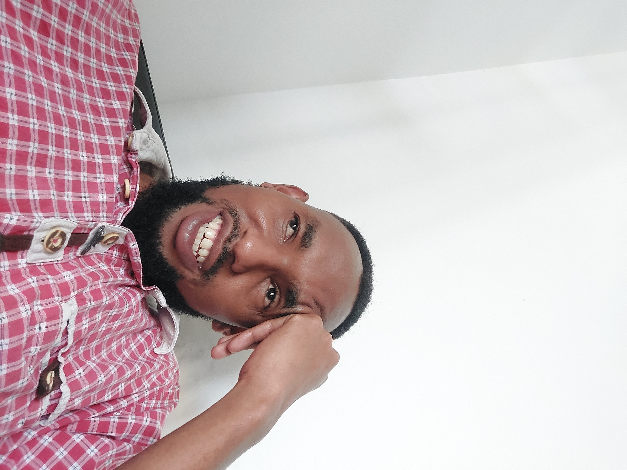A medium shot depicts a dark-skinned man with a trimmed beard and short hair, wearing a red and white checkered shirt with ornamental buttons. He is smiling, his teeth visible, with his right hand near his temple. The background is a plain white wall. The image lacks discernible features to conclusively ascertain the location as Dar es-Salaam, Tanzania. The scene is indoors with the subject presumably seated or lying down.