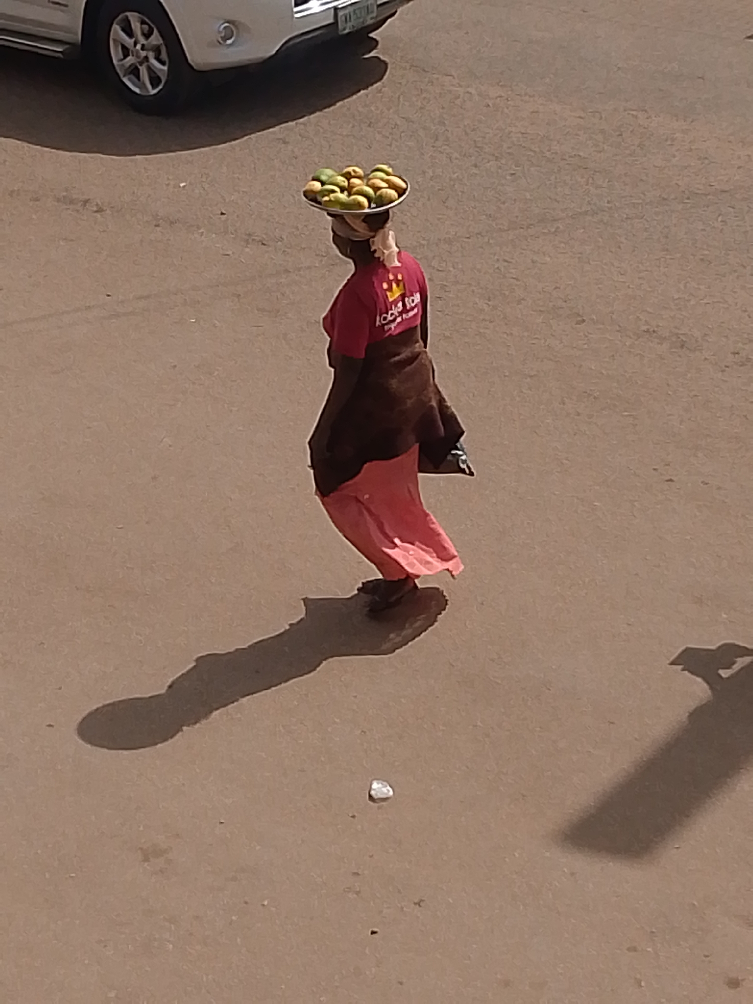 A woman is walking on a paved road in Bukuru, Nigeria. She is carrying a flat tray filled with what appears to be mangoes on her head. She is wearing a red t-shirt with text on the back, a brown wrap, and a long, coral-colored skirt. A white car is visible in the upper left corner of the image. There is trash on the ground. The scene is outdoors and appears to be during the day.
