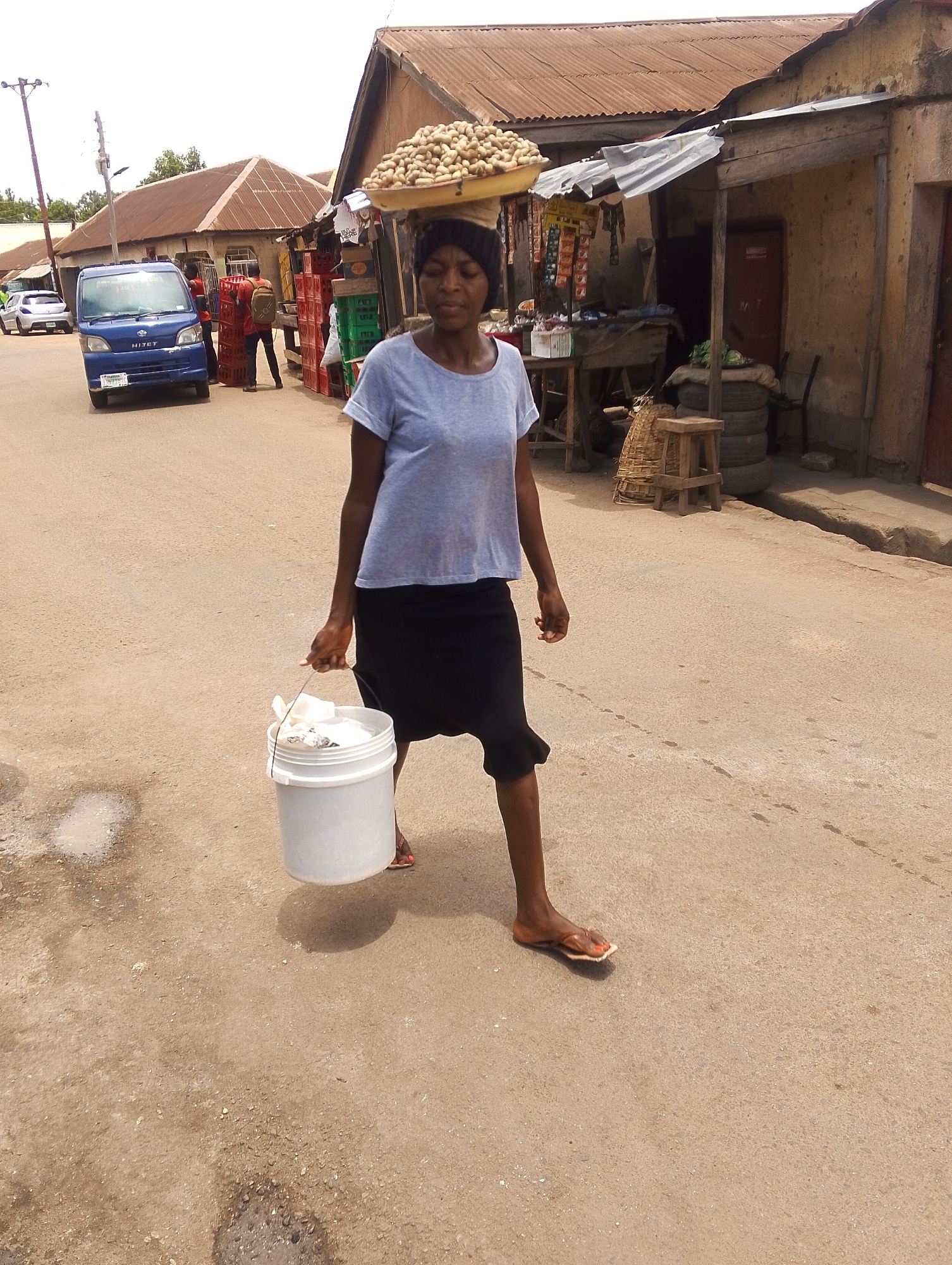 A woman is walking down a street in Jos, Nigeria. She is carrying a tray of potatoes on her head and a white bucket in her right hand. She is wearing a gray t-shirt, a black skirt, and sandals. The street is dusty and there are buildings on either side. A blue van is parked on the left side of the street. There are other people in the background. The sky is overcast.