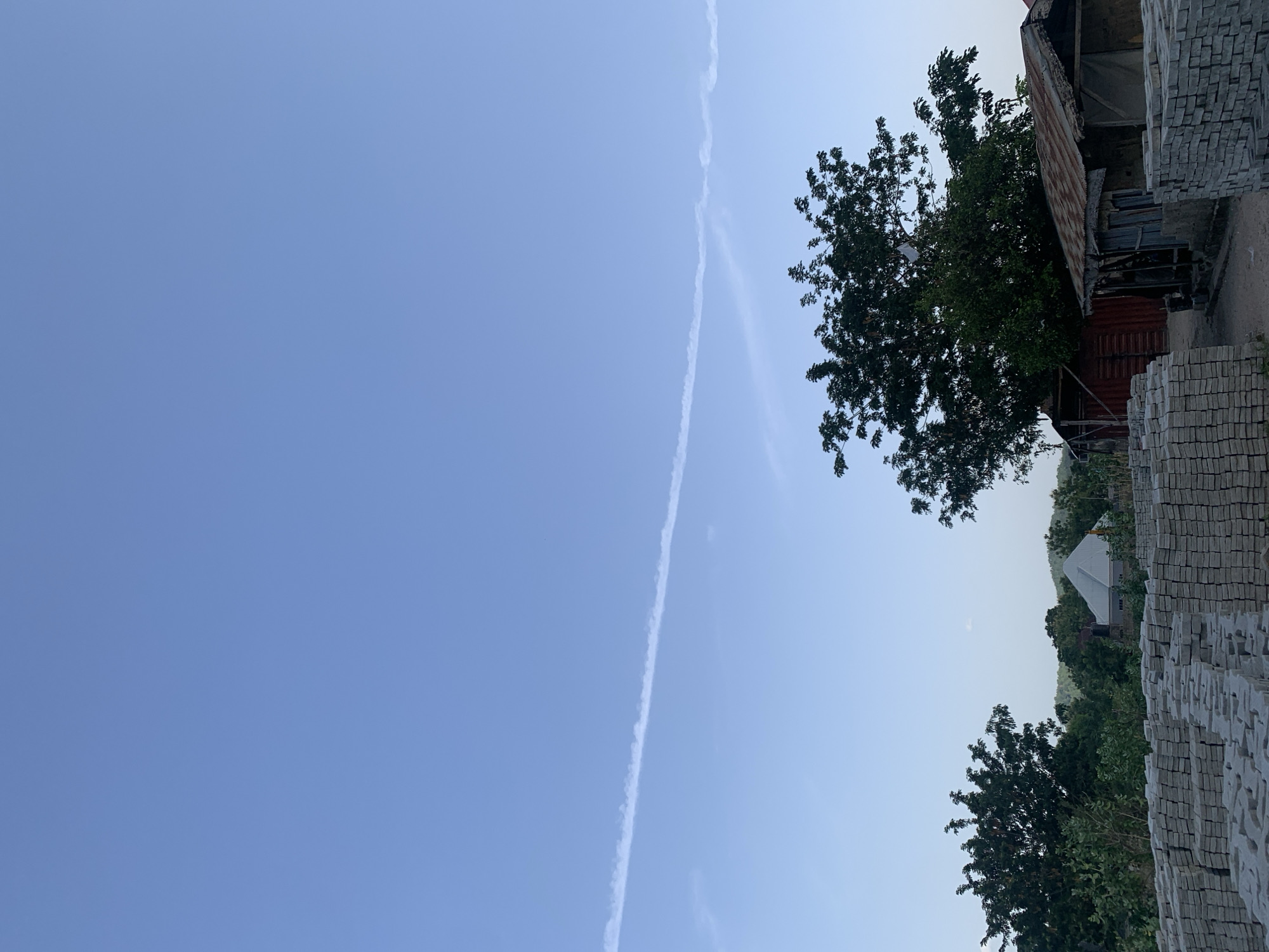 The image shows a clear blue sky with a contrail stretching across it. In the foreground, there are stacks of light gray bricks. A tree stands near the bricks, partially obscuring a dilapidated structure with a rusty corrugated metal roof. In the distance, a white-roofed building is visible among the trees. The photo appears to be taken in Zango-ndaji, Nigeria. No people are visible in the frame.