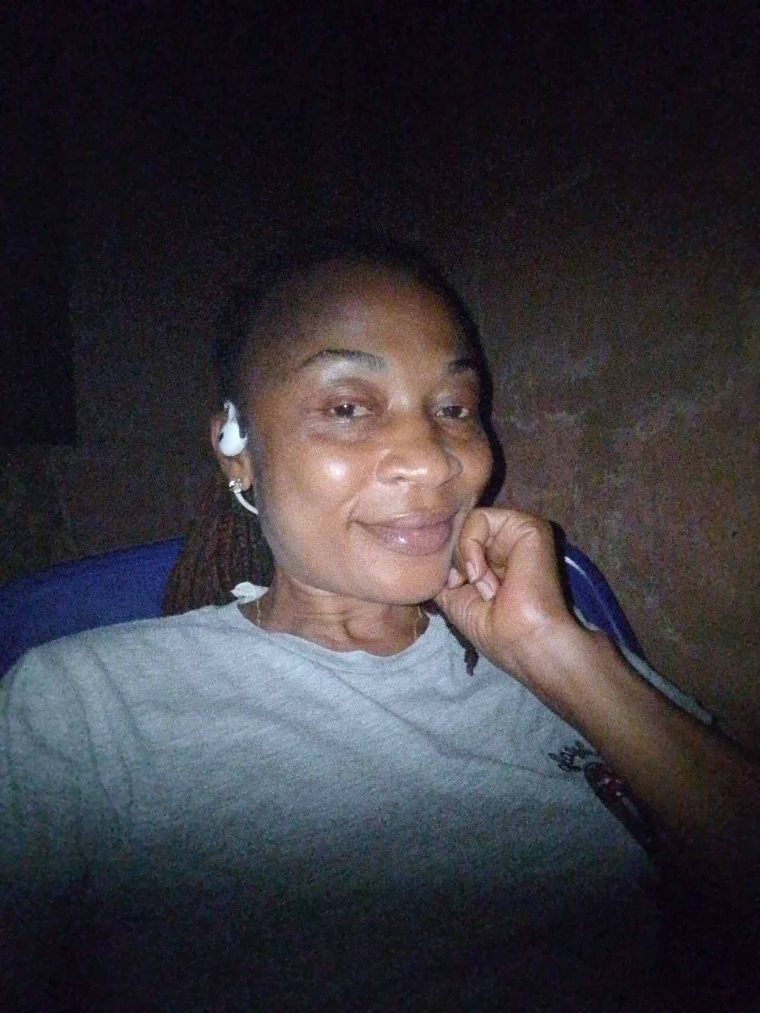 A selfie shows a woman in a dimly lit environment. She is looking at the camera and smiling. She is wearing a gray t-shirt and has white earbuds in her ears. Her right hand is resting on her chin. The background appears to be a wall. The image was likely taken in Aka, Nigeria.