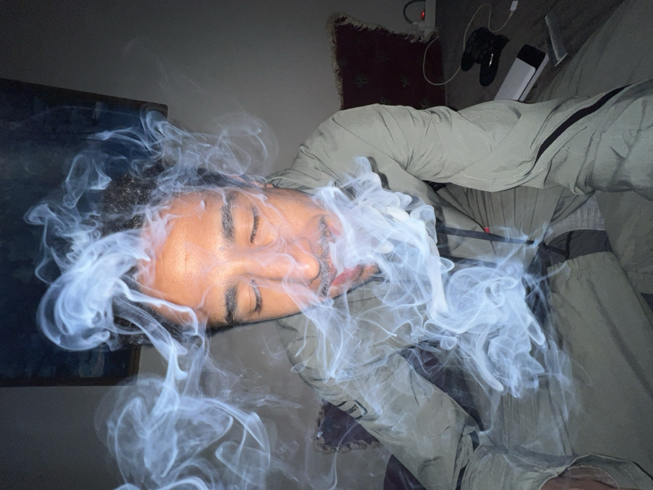 An individual is depicted indoors, in what appears to be a residential setting within the city of Tanger, Morocco. The subject, a male with dark curly hair and a mustache, is seated and positioned centrally in the frame, facing slightly downwards with closed eyes. He wears a light olive-green or khaki two-piece garment, possibly a tracksuit, featuring dark vertical stripes on the sleeves and legs. A faint patch on the left sleeve displays text, possibly "MARMOT." A significant volume of light-colored smoke envelops the individual's head and upper torso, swirling upwards and outwards from the chest and face area.
In the background, on either side of the individual, dark red patterned cushions or pillows are visible. A dark-framed object, likely a picture or mirror, is partially visible on the wall behind the subject's head. To the right of the individual, on a surface resembling a floor or low table, several objects are arranged: a black item consistent with a controller or glove, a white portable power bank, a white charging block or adapter plugged into a socket with a small orange indicator light, and an associated cable. The room is dimly lit, with light-colored walls fading into darkness at the top. The scene suggests a calm or contemplative atmosphere.