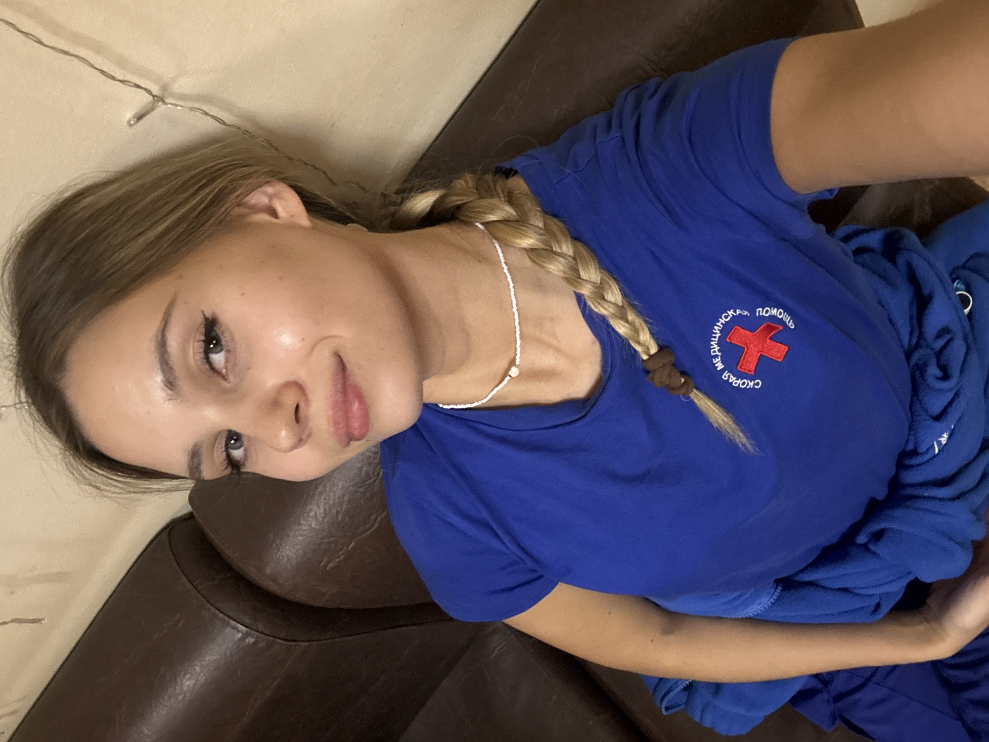 A young woman with blonde hair braided and secured with a brown tie is pictured in a close-up, selfie-style shot. She is wearing a blue t-shirt with Russian writing and a red cross emblem. She is wearing a white beaded necklace. She is reclining on a dark brown leather sofa. A strand of lights can be seen near the top left of the frame. The location is likely Chaikovsky, Russia, based on the writing on the t-shirt.