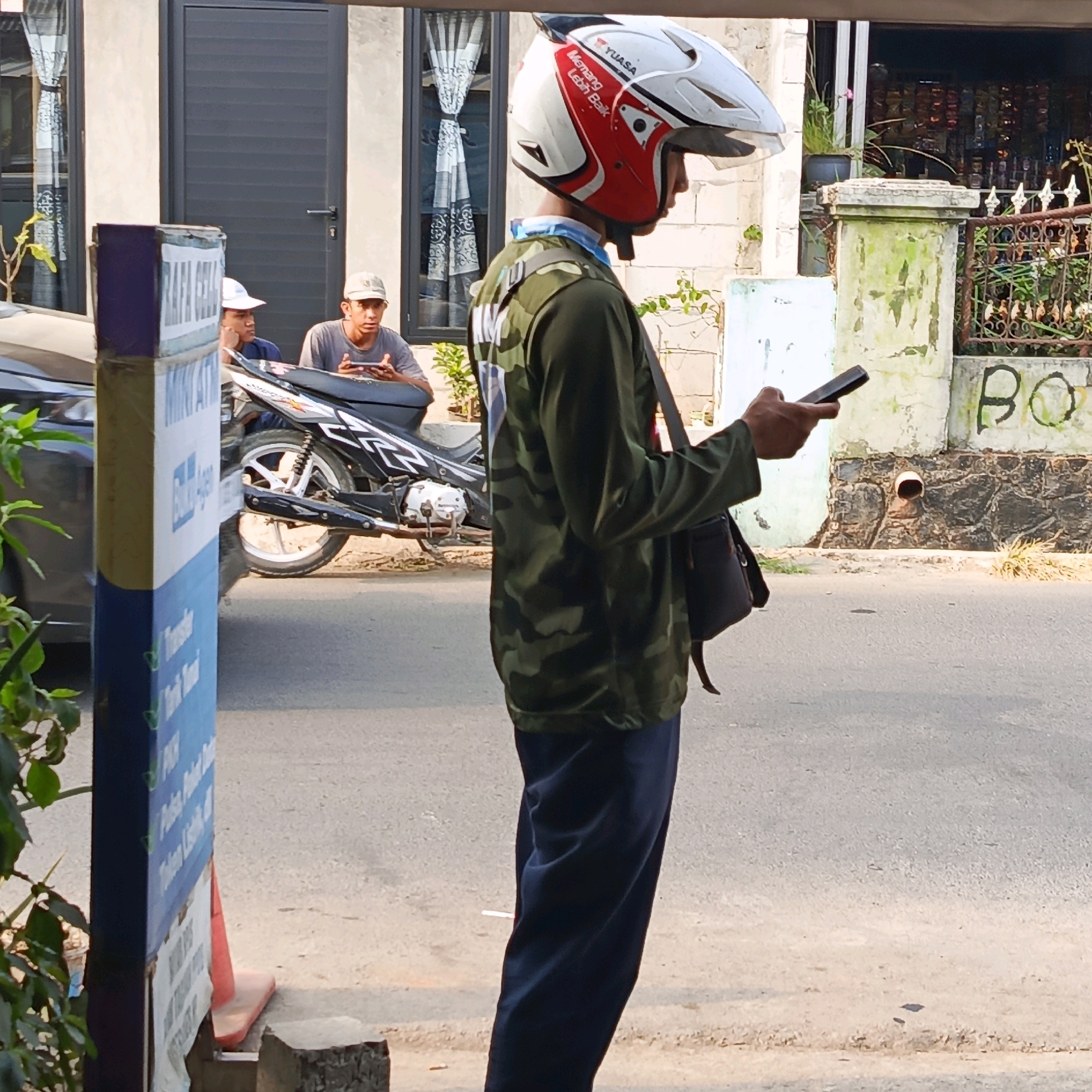 A man wearing a camouflage jacket and a white and red helmet is standing on a sidewalk, looking at his phone. He has a dark bag slung over his shoulder. In the background, two other men are sitting on a motorcycle. One of them is also looking at his phone. The scene appears to be on a street in Karawang, Indonesia, with a building and a signpost visible.