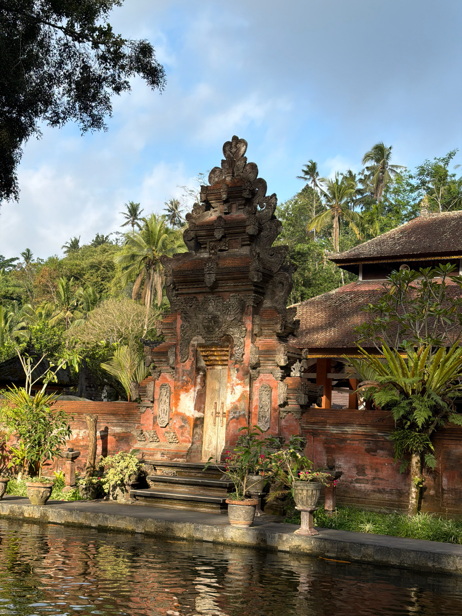 This image shows an outdoor view of the Tirta Empul Temple in Tampaksiring, Indonesia. In the foreground, a body of water reflects the surrounding environment. To the right of the water is a stone pathway and steps leading up to a structure. The structure features red brick walls, ornate carvings, and a tiled roof. A large, multi-tiered gateway with intricate stone carvings stands prominently in the center, with a wooden door. Several potted plants, including ferns and flowering plants, are placed near the steps and along the pathway. Behind the temple structures, lush green vegetation and palm trees cover the rolling hills. The sky above is blue with scattered clouds. The image appears to have been taken during daylight.