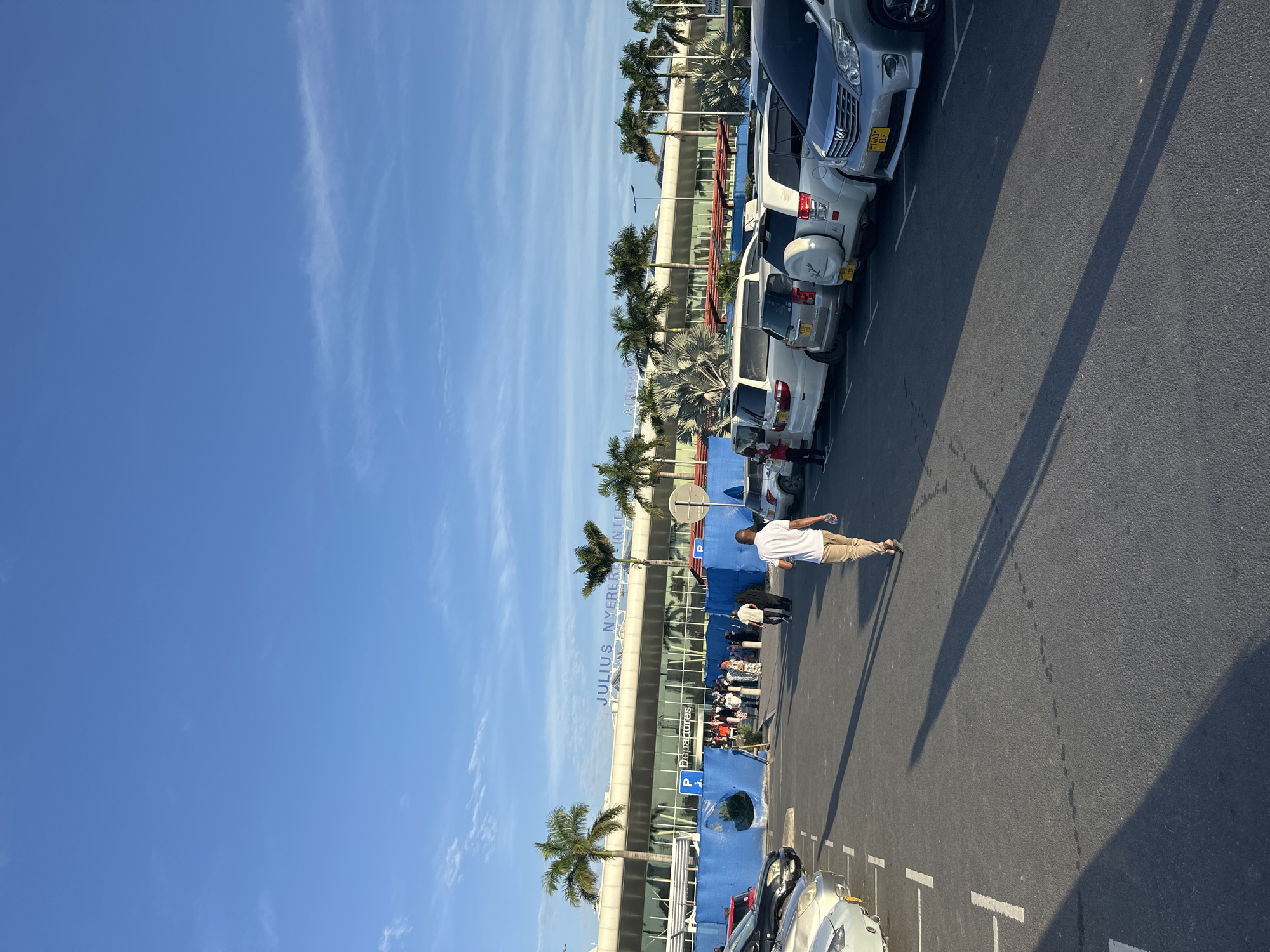 The image depicts Julius Nyerere International Airport in Dar es Salaam, Tanzania. The view is an eye-level shot of the exterior of the airport building, taken from the parking area. The bright blue sky with thin cloud streaks dominates the upper portion of the image. Palm trees line the front of the airport building. People are seen entering and exiting the airport; some are standing in groups. Cars are parked on the right side of the frame, along with parking space markings painted in white. A man in a white shirt and khaki pants is walking across the parking area. Shadows are cast on the ground from the sun.