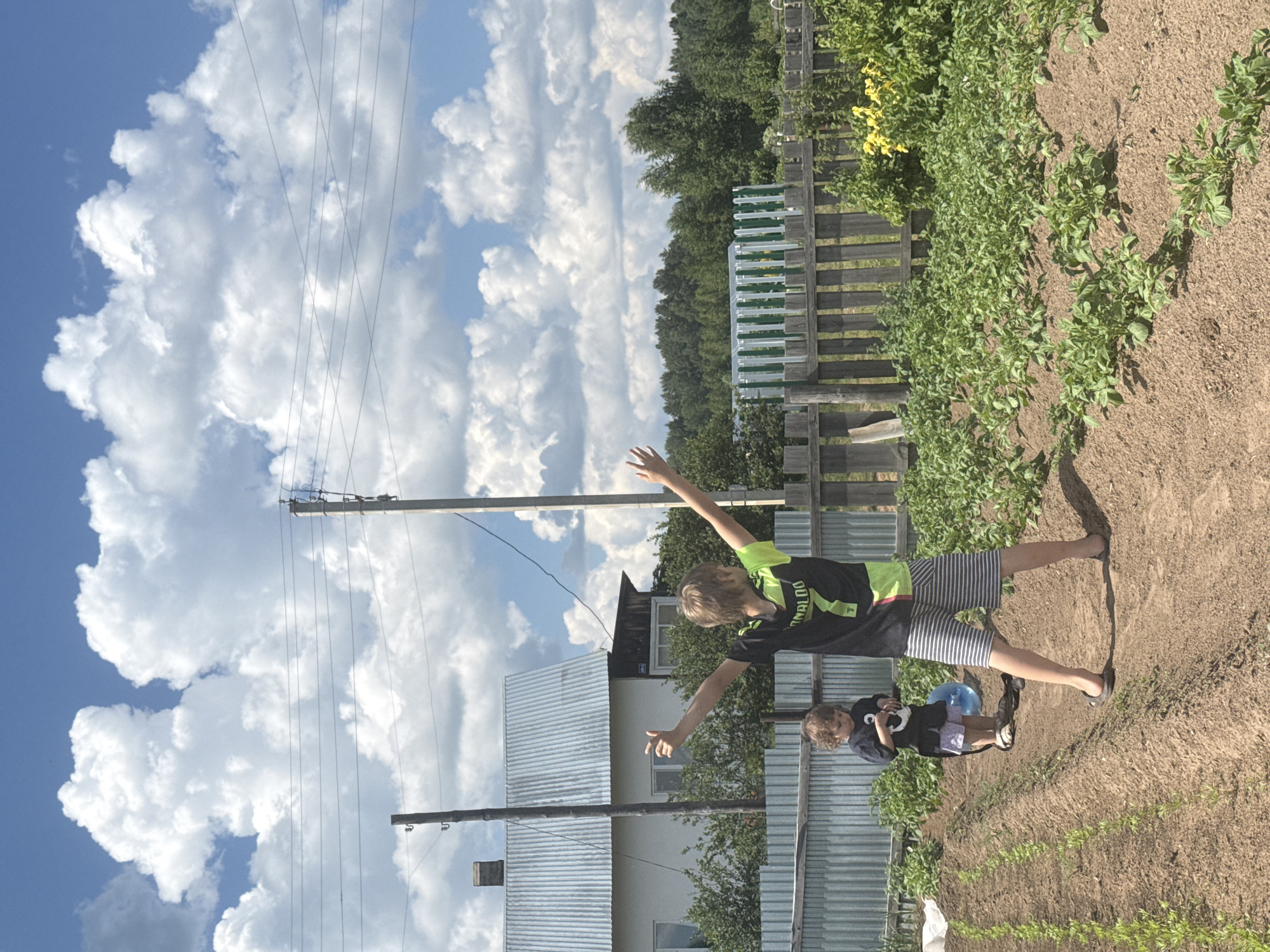 In a rural outdoor setting, likely a vegetable garden, two individuals are present on a sunny day with a blue sky and fluffy white clouds. The first individual, a young boy wearing a black t-shirt with "RONALDO 7" and lime green accents, and striped shorts, stands with his arms raised to the sides, facing away from the camera. The second individual, a young child in a dark t-shirt and lighter shorts, stands to the left and slightly in front of the boy, holding a blue object. They are positioned in a garden with rows of tilled soil and green plants. In the background, a wooden fence separates the garden from a building with a corrugated metal roof and a green and white fenced greenhouse. Trees and power lines are also visible in the distance. The location is identified as Ural'skoye, Russia.