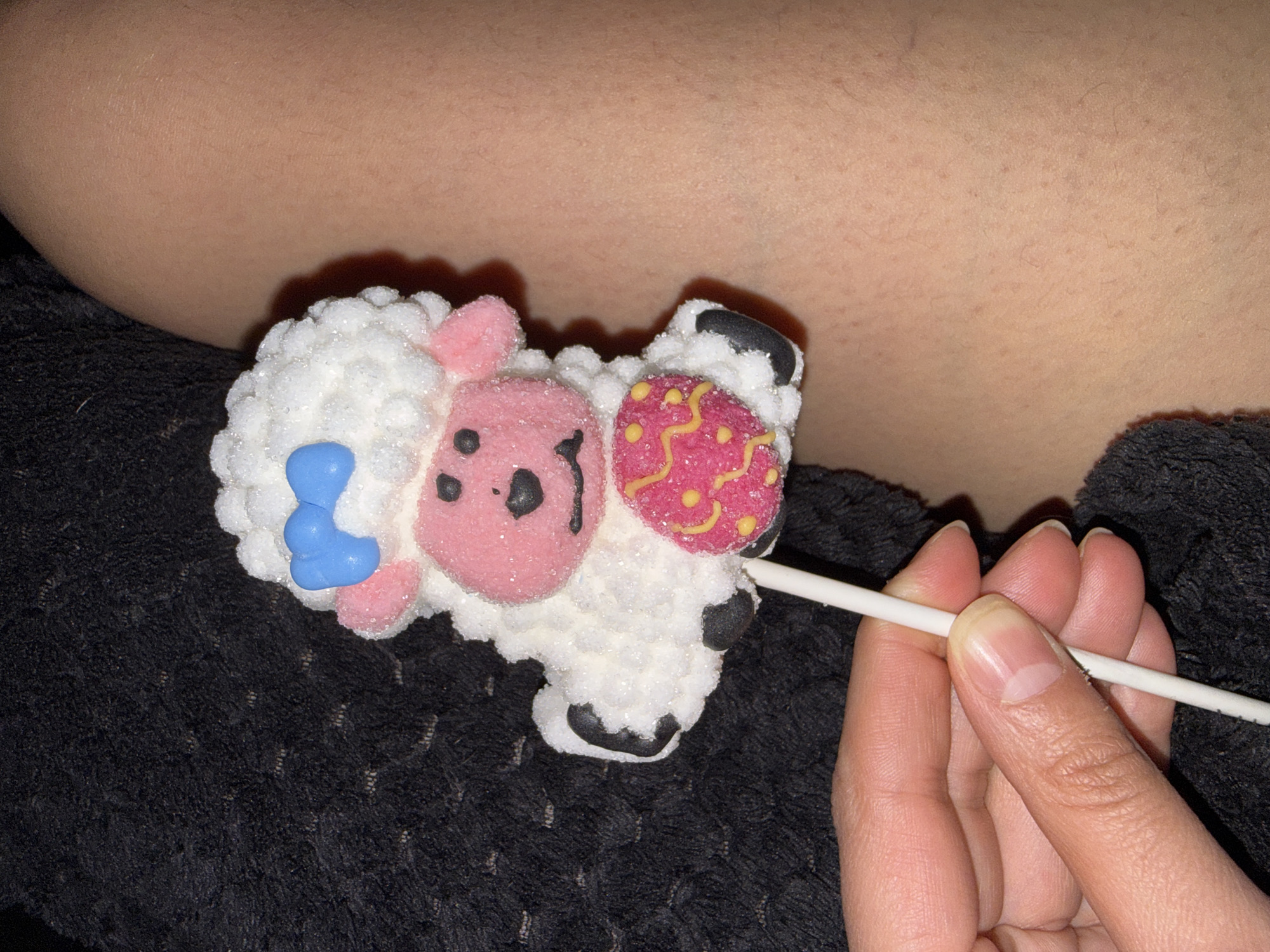 A close-up shot depicts a person holding a lamb-shaped marshmallow lollipop. The lamb is primarily white with a textured, sugar-like coating, adorned with pink facial features and a blue bow. A decorated pink egg-shaped detail is attached to the side of the lamb. The lollipop is held with a stick in the person's hand. The background consists of a person's bare skin and a dark, textured fabric. There is no evidence to suggest the location is in Great Yarmouth, United Kingdom.