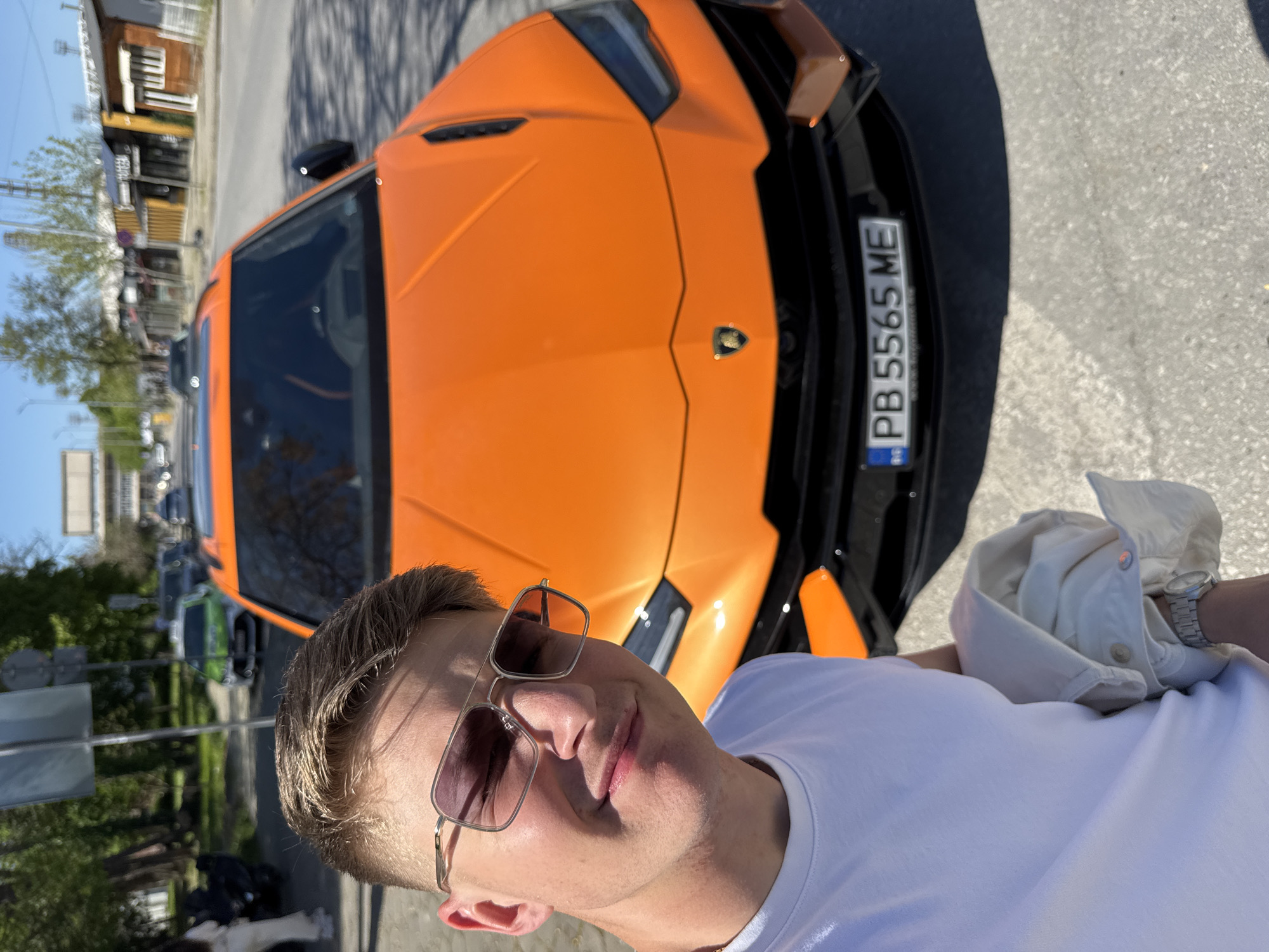 A man in a white t-shirt and sunglasses is standing next to an orange Lamborghini Urus with a Bulgarian license plate reading "PB 5565 ME". The location appears to be a street in Plovdiv, Bulgaria, suggested by the license plate and architectural style. The environment includes a city street, trees, and buildings in the background. The man is posing for a selfie.