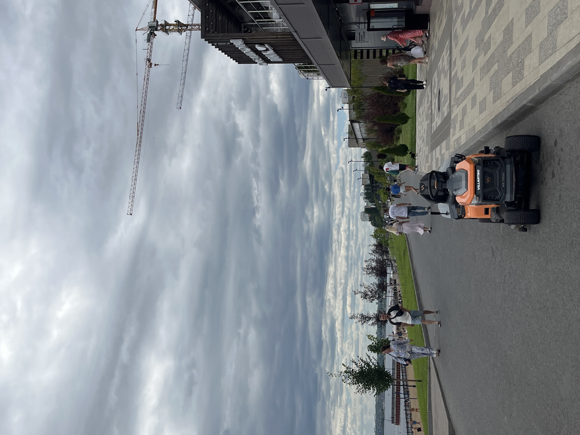 The image depicts a cloudy day in Saratov, Russia, specifically on the embankment of the Volga River. In the foreground, an orange riding lawnmower is parked on the asphalt road. Several people are walking along the paved pathway adjacent to the road. On the left side of the image, a grassy area with trees leads down to the river, where people can be seen on a beach. The background features modern buildings and construction cranes, indicating ongoing development. The overall scene is one of urban life on a leisurely day by the water.
