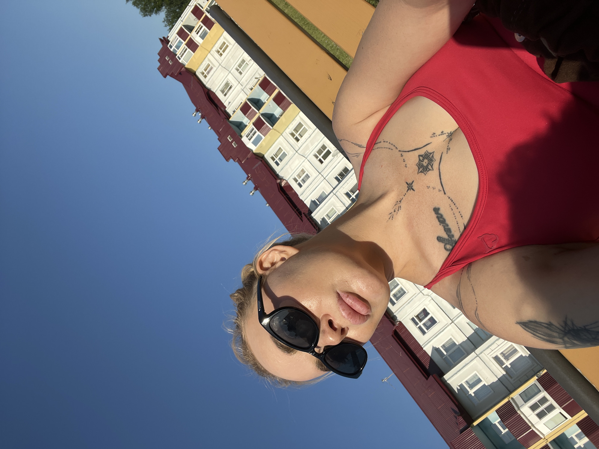 A blonde woman with sunglasses and a red top poses for a selfie on a park bench with a building in the background. The building is multi-story with a red roof and yellow and white siding. The sky is clear and blue. The woman has tattoos on her chest and arm.