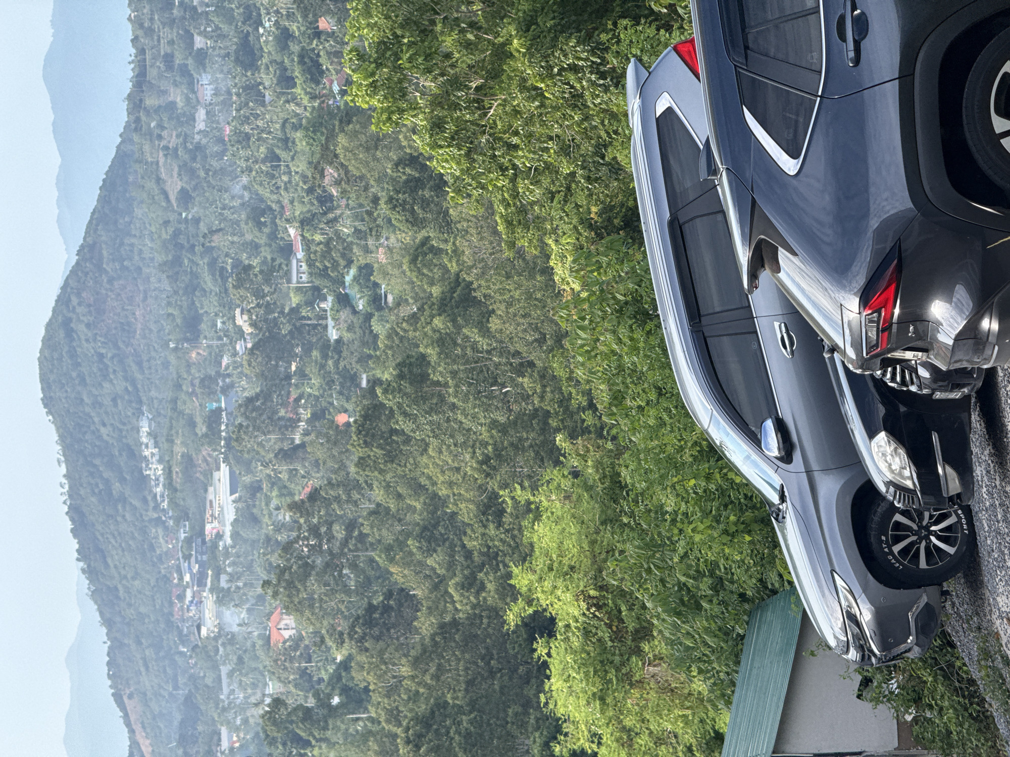 In Ko Samui District, Thailand, on a hazy day, a row of cars is parked on a gravel incline. The foreground features the rear of a dark gray SUV, its taillights illuminated. To its left, a silver SUV is parked, with its front fender and wheel visible. Further left, a black sedan is partially obscured. Beyond the vehicles, a dense, green forest covers the landscape, leading up to rolling hills dotted with buildings and houses. The sky is a pale blue, with faint mountain ranges in the distance.
