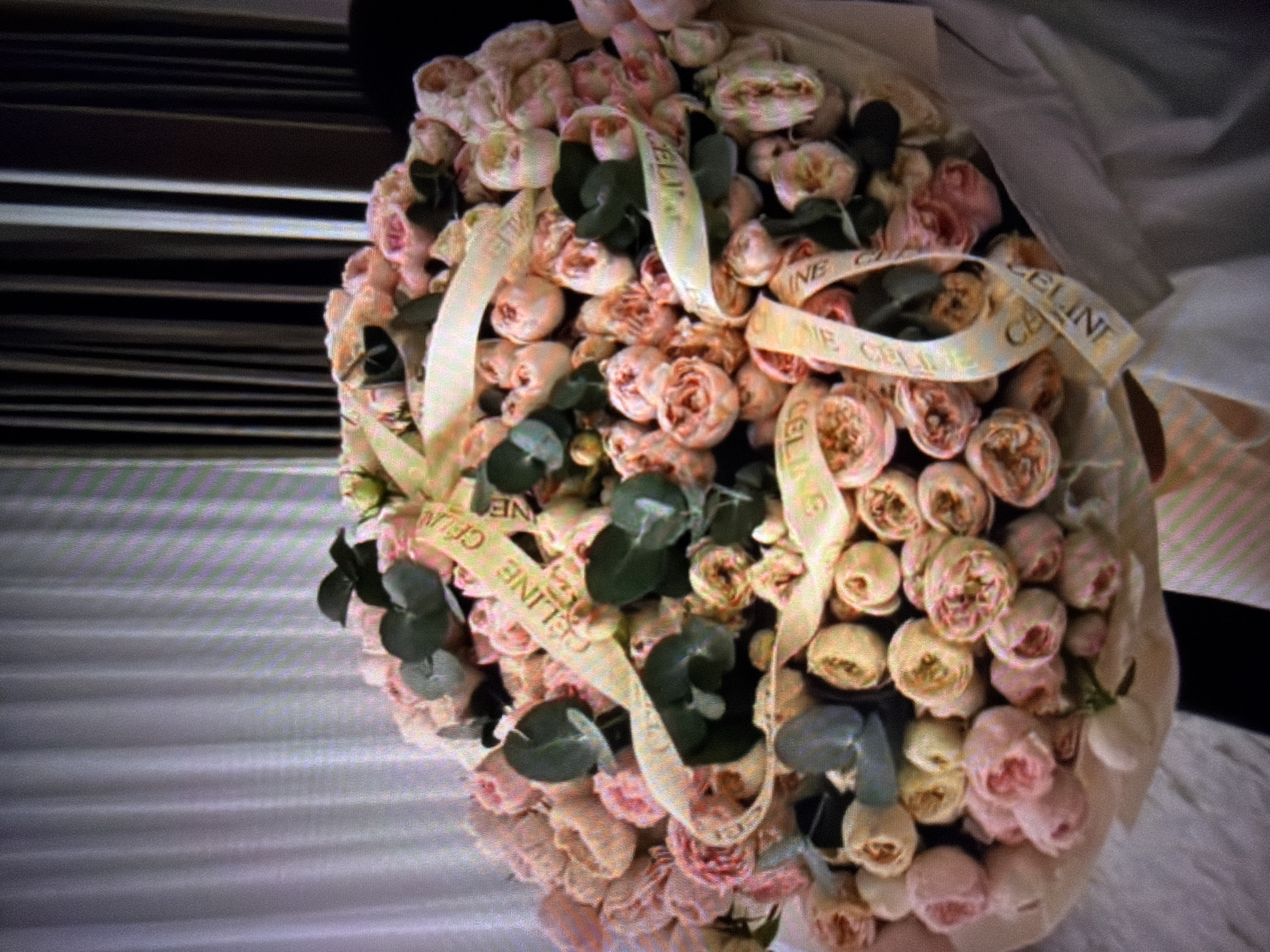 A bouquet of light pink and cream roses with dark green leaves is tied with a light yellow ribbon with the word "CELINE" repeated in a black font. The bouquet is placed on a light-colored surface, possibly a table or a wrapped gift. In the background, there are vertical blinds that are light gray on the left and dark gray on the right. The image is a close-up shot of the bouquet, with the background slightly blurred. There is no indication of the location being in Beșalma, Moldova.