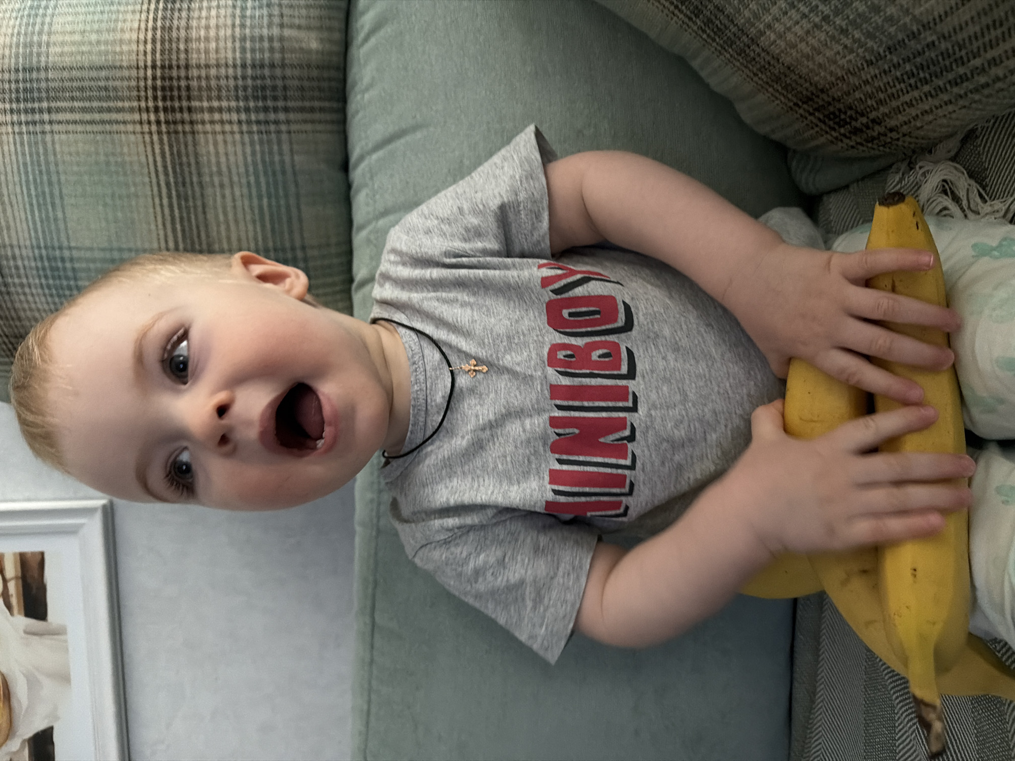 A toddler wearing a gray t-shirt with "MINI BOY" in red lettering is lying on a couch, holding a bunch of bananas. The couch is upholstered in a light green fabric and has a plaid cushion. There is a picture frame in the bottom left corner of the image. The location is not identified as Votkinsk, Russia.