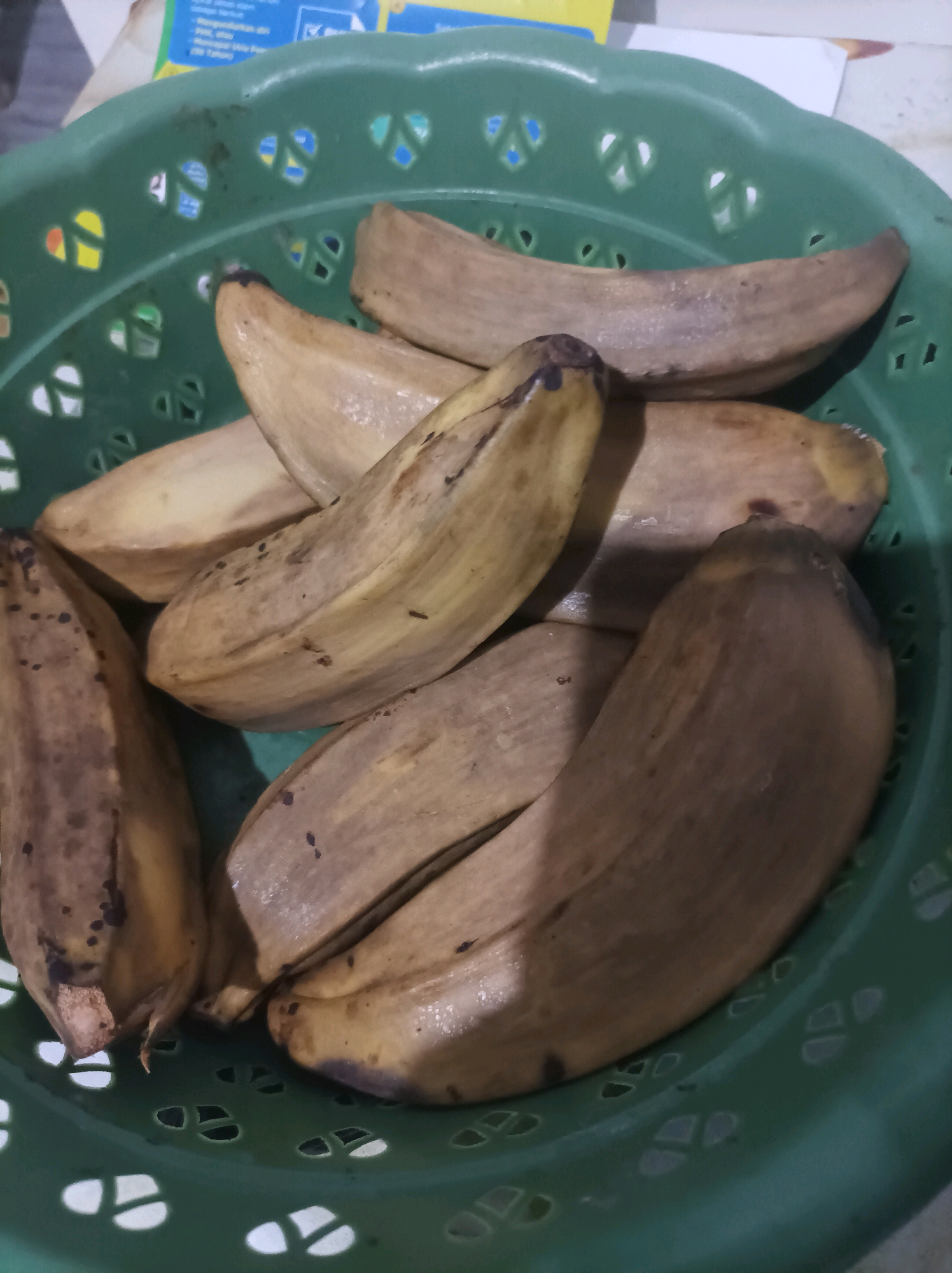 A close-up shot captures a green plastic basket filled with a pile of peeled bananas. The bananas are mostly light brown with some darker brown spots and blemishes, indicating ripeness. The basket's surface has a patterned design with open cutouts. The background is blurred, showing hints of paper and a colorful object. The scene appears to be indoors, likely in a kitchen or storage area, in the city of Canga'an, Indonesia.