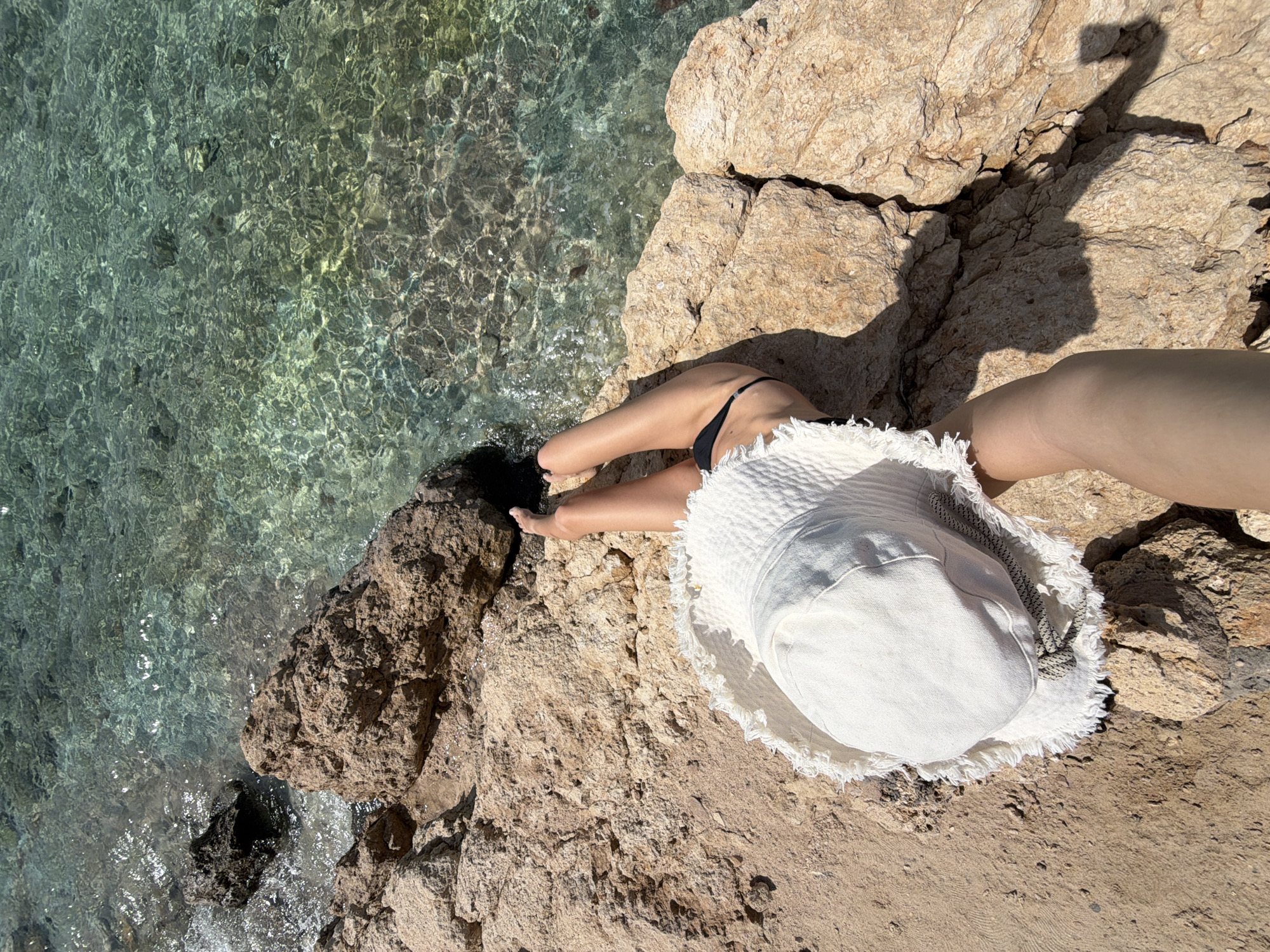 A high-angle shot shows a sunbather at the edge of clear turquoise water, likely the Red Sea, near a rocky coastline, possibly near Sharm El Sheikh, Egypt. The person is wearing a black swimsuit bottom, sitting on a tan rock formation. A white floppy hat with frayed edges lies beside them. The water is clear enough to see the rocky bottom. The lighting suggests it is a sunny day, and there is a shadow of a hand holding a phone.