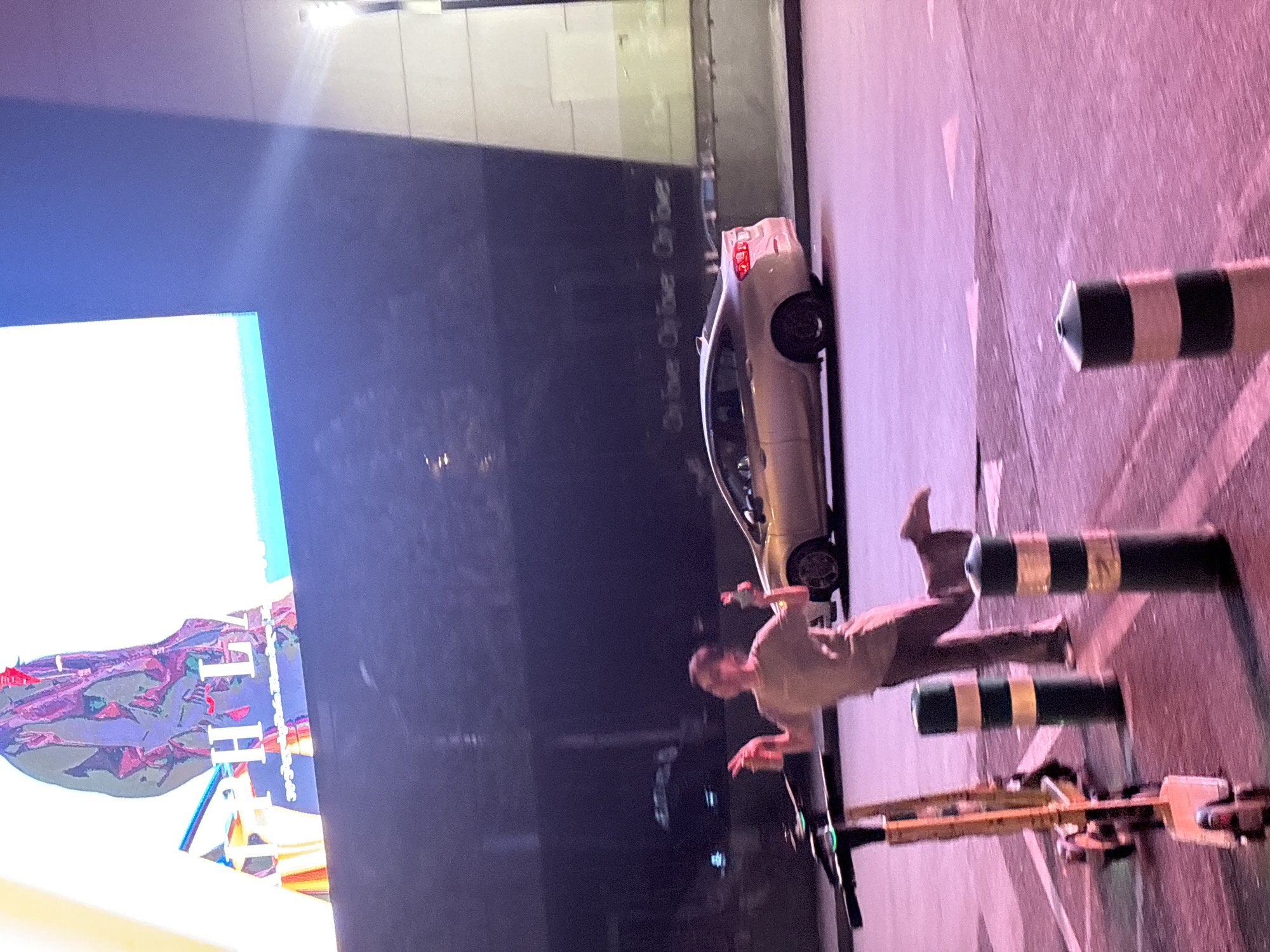 In Tbilisi, Georgia, at night, a person is captured mid-stride on a street, appearing to be dancing or posing. They are wearing a tan t-shirt and pants and are holding a bottle in each hand, with their arms raised. The person is positioned near several parked electric scooters and bollards, with a silver sedan driving past in the background. Above, a large digital billboard displays an abstract image with white lettering, partially visible. The scene is illuminated by the glow of the billboard and streetlights, casting a purplish hue on the pavement.