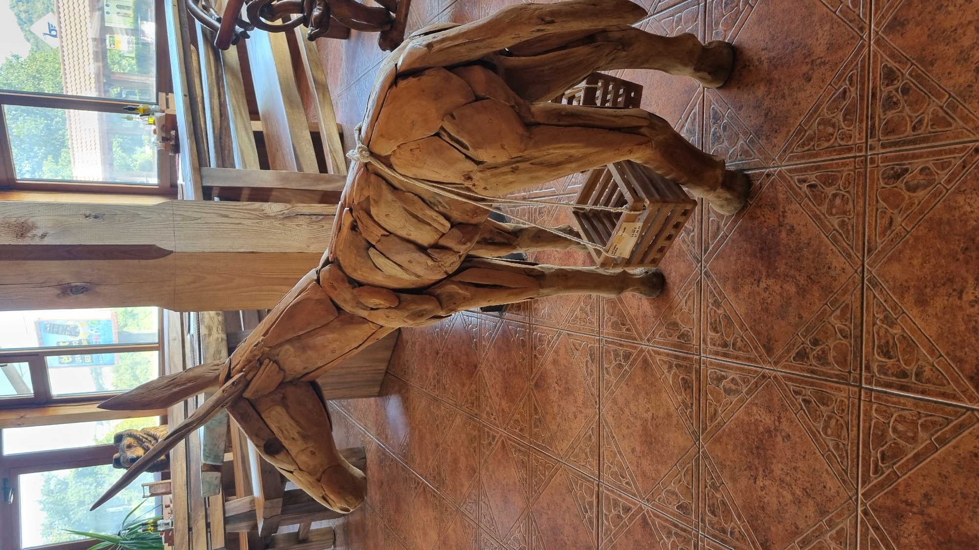 The image depicts a life-size wooden sculpture of a donkey, crafted from what appears to be driftwood or similarly shaped pieces of wood. The donkey sculpture is positioned indoors, on a tiled floor with a reddish-brown, patterned design. The donkey has a crate hanging from each side, attached with rope. In the background, large windows offer a view of an outdoor environment with greenery and buildings. The interior appears to be a rustic establishment with wooden tables and chairs visible through the windows and to the left of the donkey. The lighting suggests daytime. The scene is located in Zlatna Panega, Bulgaria, likely at a restaurant or guesthouse. There are no people or animals visible in the image, and no active interactions are occurring. The focus is entirely on the wooden donkey sculpture and its immediate surroundings.