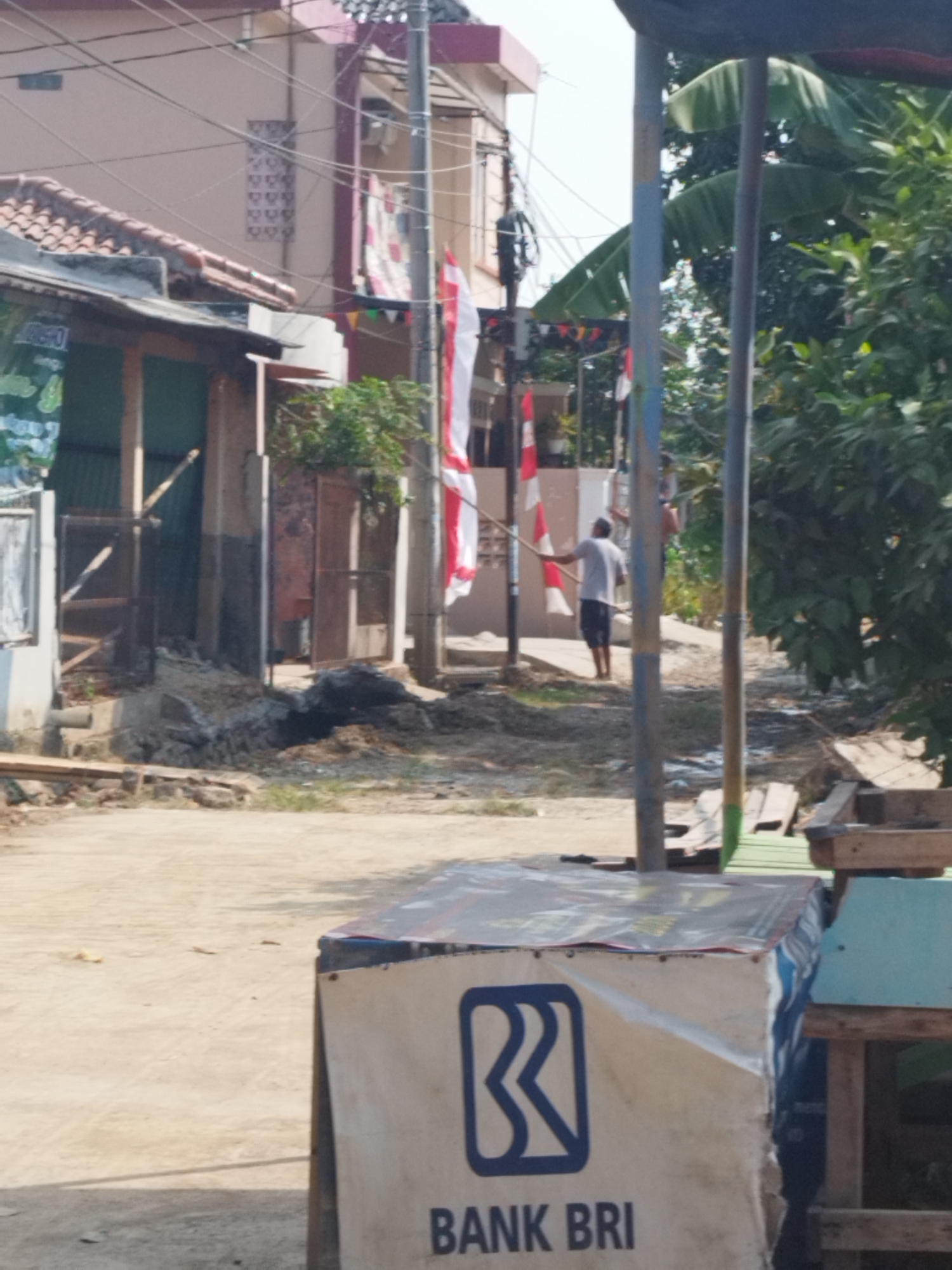The image depicts an outdoor scene in a neighborhood that appears to be undergoing some construction or renovation. In the foreground, a table covered with a white banner displaying the blue "R" logo and "BANK BRI" text is positioned. Behind this, a dirt road is visible, leading towards several buildings. One building, partially obscured by a tree, is a light-colored structure with red-tiled roofing. Adjacent to it is another building with a darker facade and a more traditional roof. In the background, two individuals are present. One person, wearing a white t-shirt and dark shorts, is standing near a pole with Indonesian flags attached, holding a long pole. Another person, wearing a light-colored shirt, is also visible in the background. The environment is characterized by green foliage from trees and banana plants. Power lines are strung across the scene. The lighting suggests it is daytime. The overall impression is a somewhat unkempt street scene in a residential area of Karawang, Indonesia.