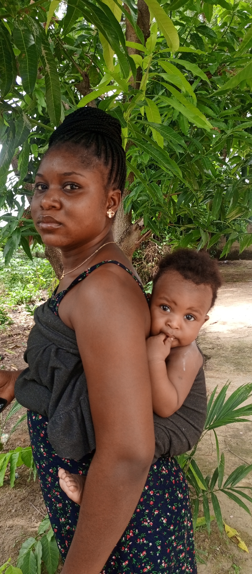 An outdoor portrait captures a woman carrying a baby on her back in Keffi, Nigeria. The woman, with dark skin and braided hair styled in an updo, wears a dark, floral-patterned dress, a thin gold-colored necklace, and a gold bow-shaped earring. She gazes directly at the camera with a neutral expression. The baby, an infant with dark skin and curly hair, is secured on her back using a dark grey fabric wrap. The baby also looks towards the camera, with a finger near its mouth and a slight drool mark on its arm, showing a curious expression. A tiny foot of the baby is visible peeking from the wrap.
The background is filled with lush green foliage, predominantly large leaves consistent with a mango tree, and a tree trunk behind the woman. The ground visible at the bottom shows earth and sparse green plants, including a small palm-like frond on the right. The scene is brightly lit by natural daylight, suggesting it is daytime. The traditional method of carrying the baby on the back is a notable cultural detail. Part of a dark object, possibly a phone, is visible in the woman's left hand. No visible text is present.