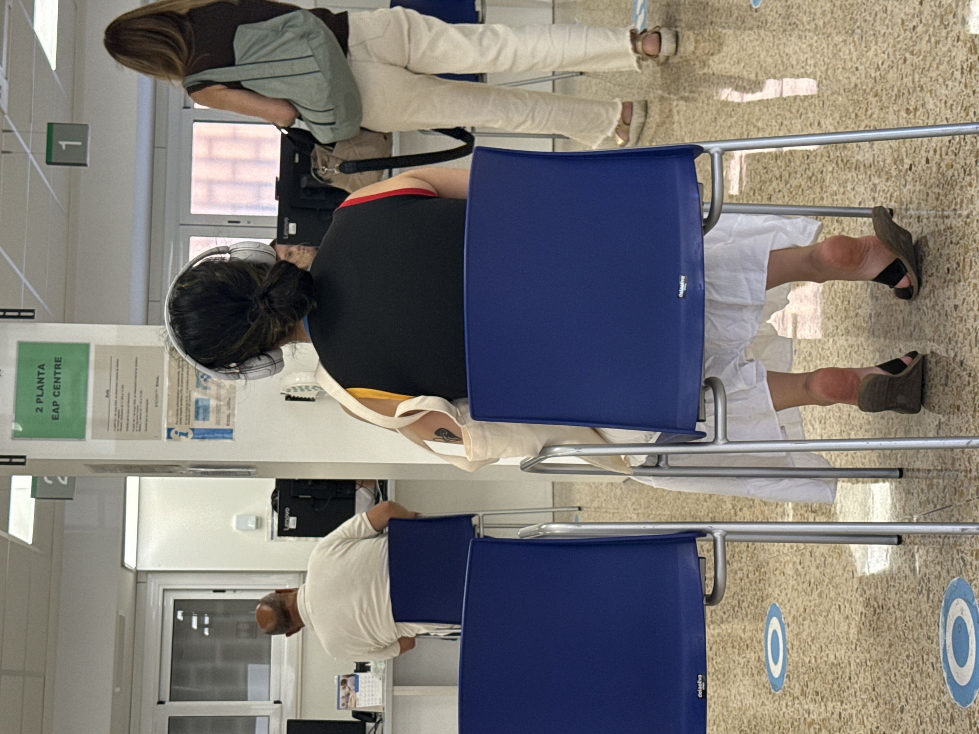 The image depicts an indoor waiting area, likely a public service center, with the signage indicating "2 PLANTA EAP CENTRE" which suggests it is the second floor of a primary care center in L'Hospitalet de Llobregat, Spain.
In the foreground, a woman with dark hair pulled back in a bun is seated, wearing white headphones and a black top with red and yellow trim. She is facing away from the camera, towards a desk area. Her feet, clad in dark sandals, are visible, and she is wearing a long white skirt. A light-colored tote bag is slung over her shoulder, and another larger bag rests on the floor beside her.
To her left, the back of a man in a white polo shirt is visible. He is also seated, facing a desk with a computer monitor. Two empty blue chairs are positioned in the foreground, one with a logo that appears to read "delsoliva." Social distancing markers, in the form of blue and white circles, are visible on the speckled floor.
In the background, another woman with long brown hair is standing, wearing white trousers and a dark top. She is carrying a light blue tote bag over her shoulder. A sign with the number "1" is mounted on the wall above her. Other desks with computer equipment and partitions are visible in the background, suggesting a functional office or service environment. The lighting appears to be from overhead fluorescent fixtures.