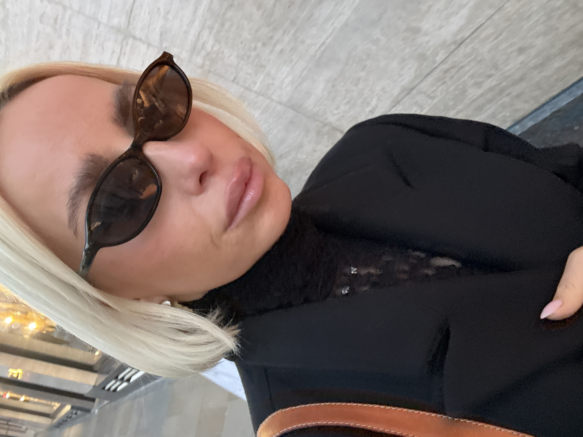 The image depicts a close-up, eye-level portrait of an individual, likely female, on a city street in London, United Kingdom. The subject has short, straight platinum blonde hair and wears dark, cat-eye sunglasses which reflect blurred urban lights and building structures. Her face shows cosmetic application, including prominent, light pink lips. She is dressed in a black blazer layered over a black turtleneck sweater. A visible layer beneath the turtleneck is a black sheer fabric, possibly lace, with two small, reflective embellishments. A tan leather strap, consistent with a shoulder bag, is visible on her left. Her right hand, displaying light pink almond-shaped manicured nails, rests on the blazer in the lower left of the frame. The background consists of a blurred, light grey, vertically textured building facade to the right, and indistinct reflections of a commercial street environment to the left.