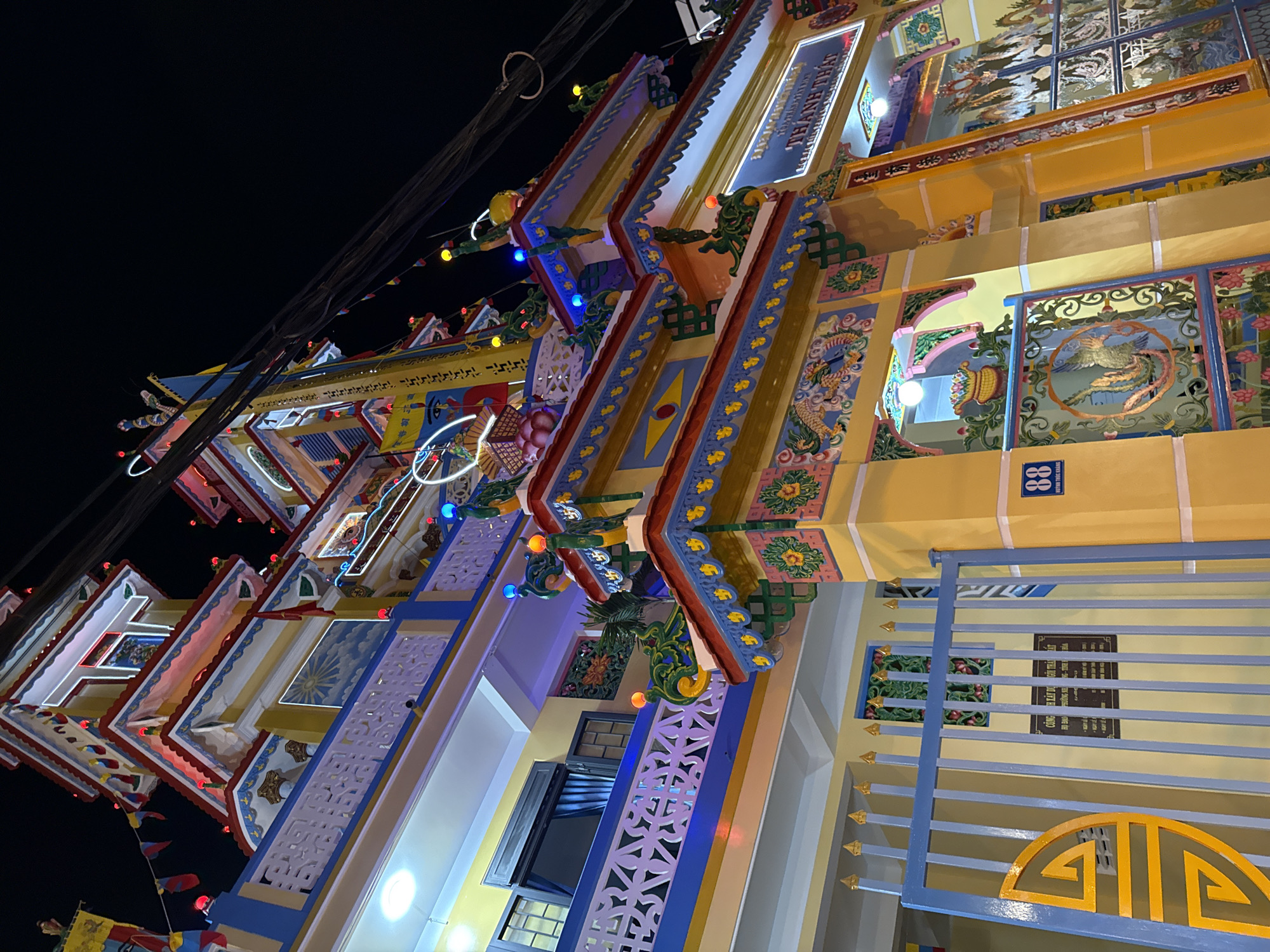 The image depicts a brightly lit, ornate temple or pagoda at night in Nha Trang, Vietnam. The structure is a multi-tiered building with a vibrant yellow exterior, adorned with intricate decorations, colorful flags, and numerous lights, including red, blue, and yellow bulbs. The architecture features a stepped roofline with decorative elements and ornate railings. A large blue gate with a golden symbol is visible on the left, leading to a pathway. Signs with Vietnamese text are posted on the wall behind the gate. The scene is dominated by the illuminated temple, creating a festive and somewhat chaotic atmosphere against the dark night sky, with visible electrical wires overhead. There are no people visible in the frame.