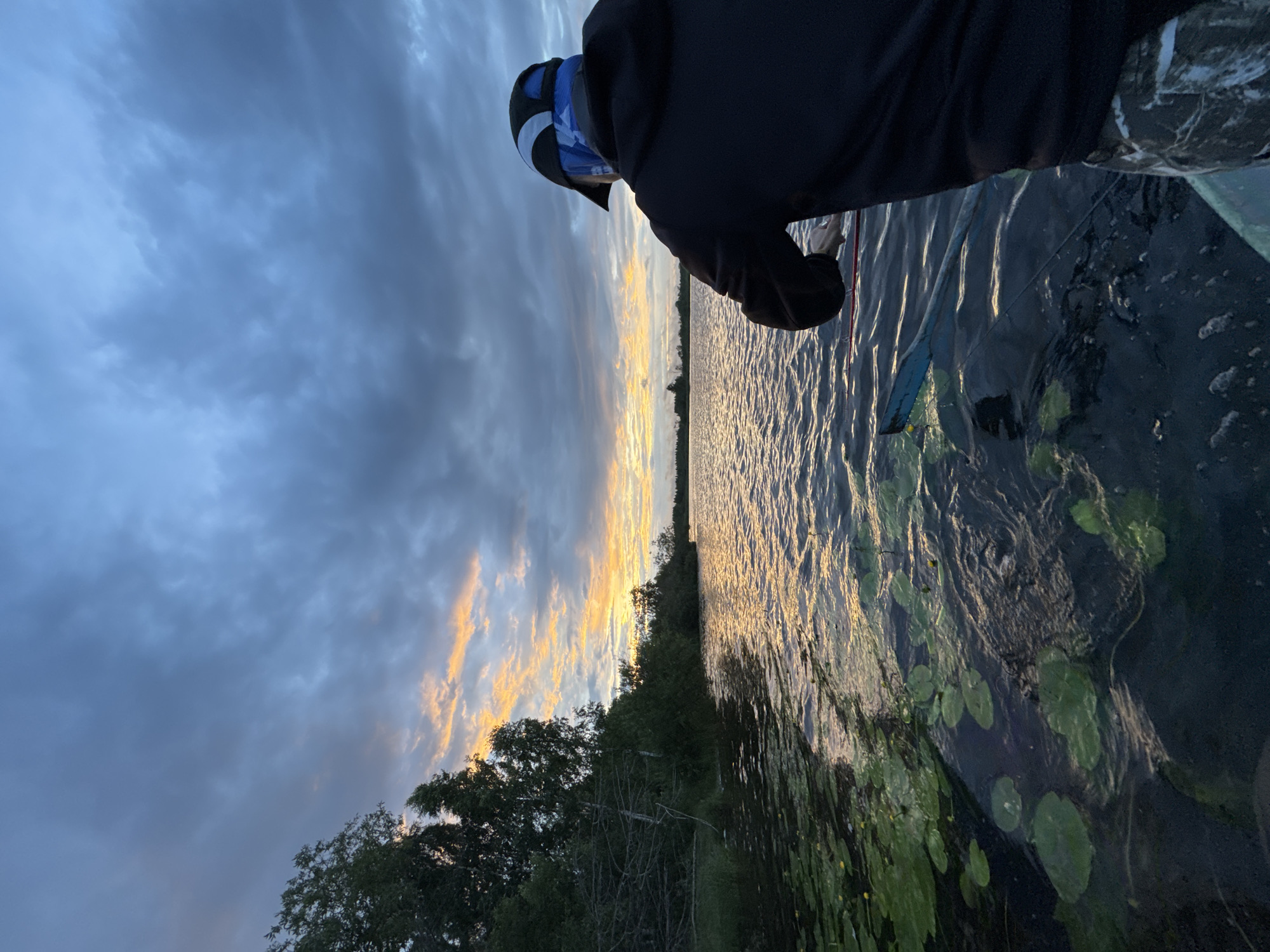 The image depicts a person in a boat on a lake at dusk. The sky is filled with dramatic clouds tinged with the orange and yellow hues of a sunset. The water is rippled, and lily pads are visible floating on the surface in the foreground. The person is wearing a black hooded sweatshirt, a baseball cap, and camouflage pants. They are holding a fishing rod. To the left, a dense line of trees and vegetation borders the lake. The scene appears to be a natural landscape, likely a rural or semi-rural area. There is no identifiable indication of the city of Saint Petersburg, Russia, in this image.