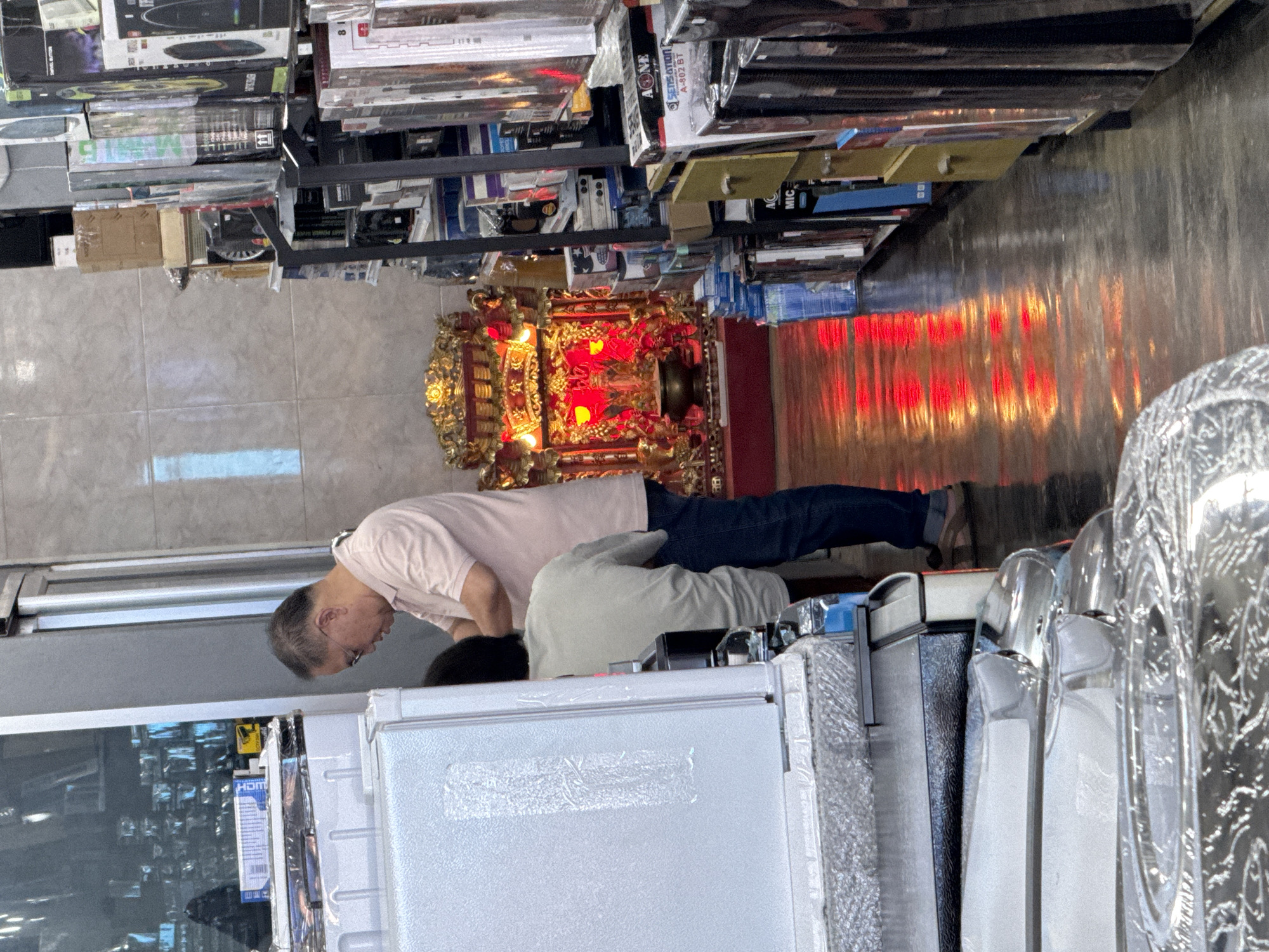 In Ranong, Thailand, a retail store is depicted with a man wearing a pink polo shirt and jeans, and another individual partially obscured from view, both standing near a display of appliances. To the right, shelves are stacked with various electronic items, and further down the aisle, a vibrant, red-lit altar is visible. The floor of the store is a reflective surface, showing a red sheen from the altar's illumination.