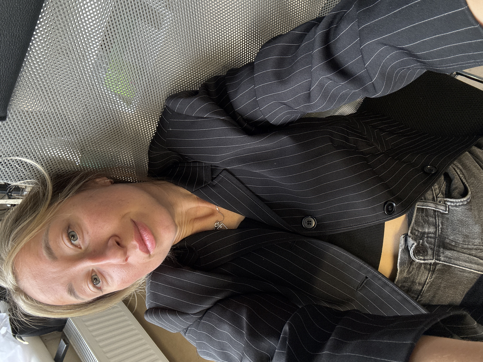 A close-up, angled shot depicts a woman with blonde hair and light eyes looking directly at the camera. She appears to be lying down or reclining in a relaxed pose. She is wearing a dark, pinstripe blazer over a dark top, with dark denim jeans visible. A silver necklace with a small pendant is around her neck.
The woman has fair skin, and her expression is neutral to slightly contemplative, with softly parted lips. Her hair is casually framing her face.
She is positioned on what appears to be a light gray, perforated mesh chair or seating. A dark, possibly leather, armrest is partially visible on the left side of the frame. Through the mesh, a subtle green object can be faintly seen in the background. In the bottom left corner, a white radiator or heating unit is partially visible, indicating an indoor setting. The overall scene suggests a casual environment, possibly an office or home, located in Chaikovsky, Russia. There is no visible text in the image.