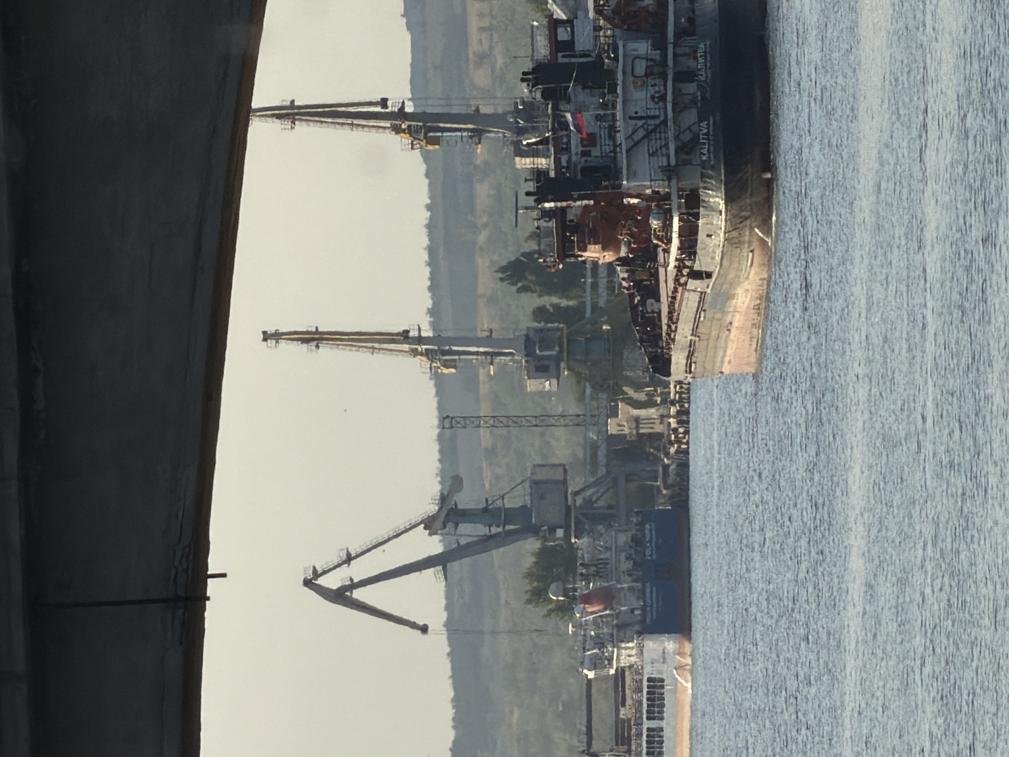 The image depicts a harbor scene in Kamyshin, Russia, with a prominent focus on maritime infrastructure and vessels. In the foreground, a large cargo ship, identified by the name "KALITVA" on its stern, sails on the water. The ship is dark-hulled with a red waterline and appears to be laden. In the mid-ground, two large harbor cranes with yellow booms are visible, suggesting cargo handling operations. One crane, a darker gray, is positioned to the left, while a lighter yellow crane stands to the right, closer to the cargo ship. Behind the cranes, a lighter-colored ship is docked, with multiple windows visible along its side. The background consists of a gently sloping, tree-covered hillside under a hazy, pale sky. A section of a dark structure, possibly a bridge or overpass, casts a shadow across the top of the image, framing the scene.