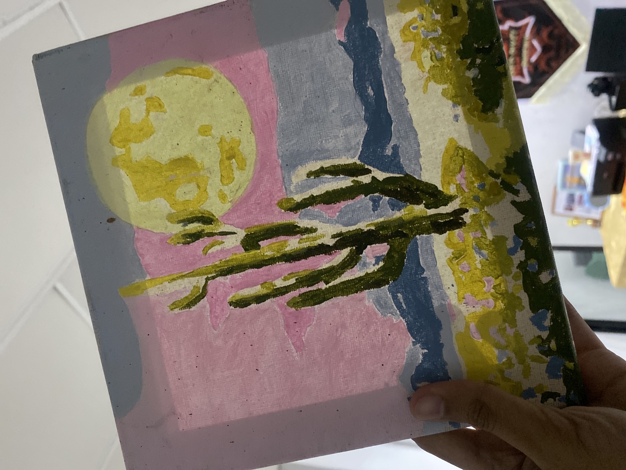 The media file primarily features a vibrant, stylized painting held by a human hand. The painting, rendered in what appears to be acrylic or gouache on canvas, depicts a landscape or natural scene with distinct blocks of color. On the left side, a large, textured yellow-green circle, suggestive of a moon or sun, stands out against a background divided into vertical bands of light blue and pink. Extending from the center, thick, dark green brushstrokes form branch-like structures or foliage. The right side of the painting transitions into an abstract area characterized by irregular patches and streaks of light green, yellow, and blue, evoking further foliage or textural elements. Visible brushstrokes and the weave of the canvas add to the painting's tactile quality.
A human hand, with a light brown skin tone, is visible holding the bottom right corner of the artwork. The background of the photograph is out of focus, revealing a white or light-colored surface. In the upper right, a dark, indistinct object is present. Below it, a reddish-brown, possibly octagonal object with some light reflections and blurry, unreadable text or a symbol is discernible. Other blurred items can be seen in the distant right background. The overall lighting suggests an indoor setting. There are no specific location cues visible within the image to indicate Davao City, Philippines.
