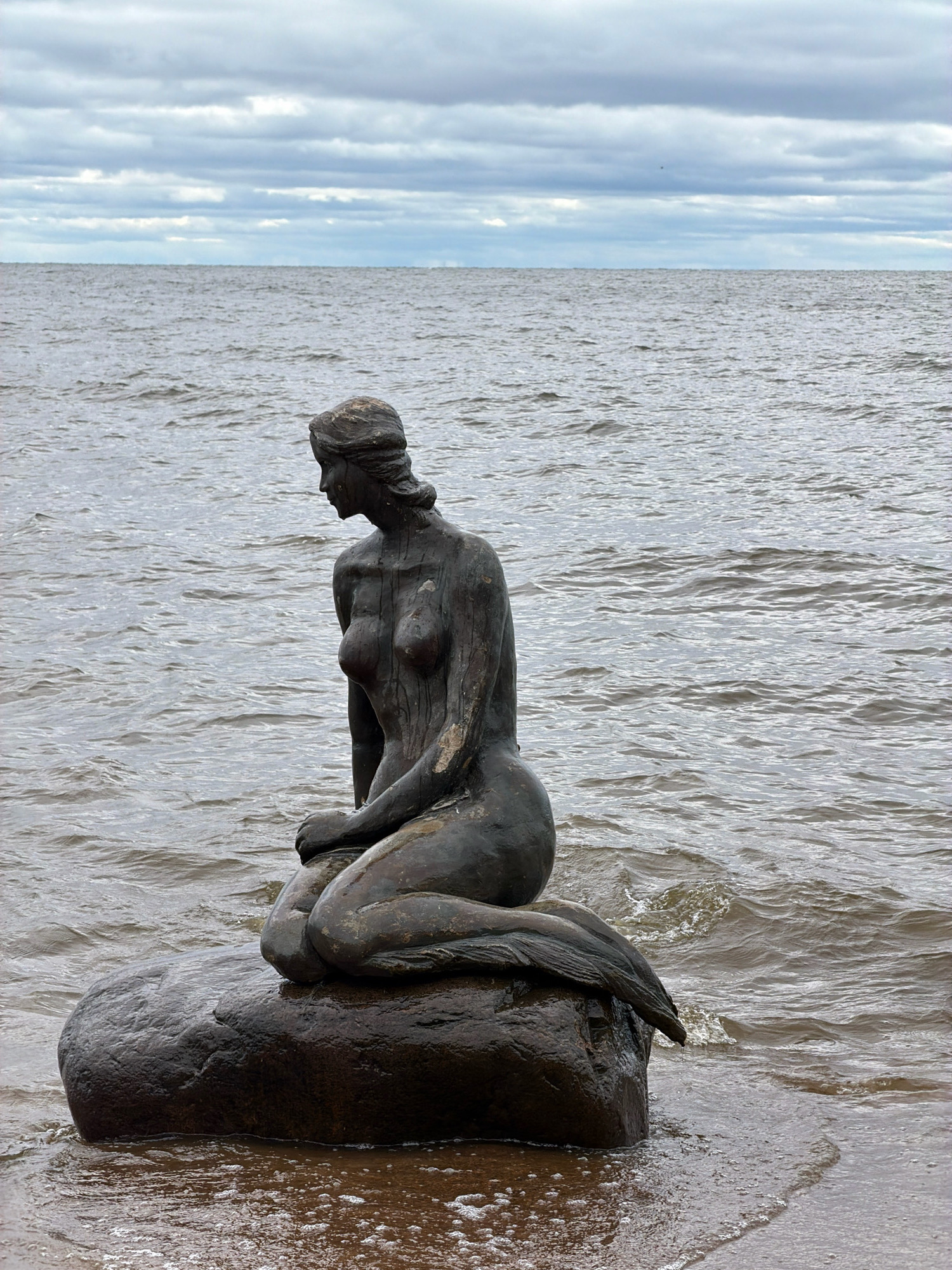 A bronze statue of a seated mermaid rests on a large, wet rock at the edge of a body of water under a cloudy sky. The mermaid is depicted with her knees drawn up, arms resting on her legs, and her head turned to her left. She appears to be wearing a band around her head. Her tail extends behind her, partially submerged in the water. The water is choppy with small waves lapping at the rock and shore. The background consists of the vast expanse of water and a heavily clouded sky. The statue is located on the shore of the Gulf of Finland in Komarovo, Russia, a coastal resort town known for its beaches and pine forests.
