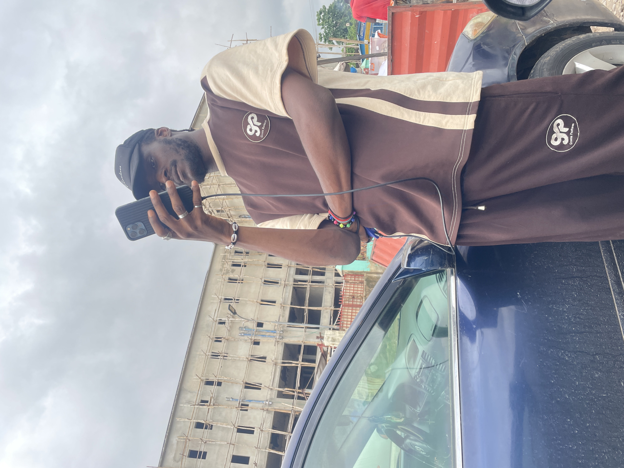 In the city of Galadimawa Village, Nigeria, a young man stands beside a blue car, holding a smartphone to take a photo of himself or the surroundings. He is wearing a brown and tan t-shirt with a logo and matching brown sweatpants. The man has a beard and is wearing a black baseball cap. In the background, a building under construction is visible, with scaffolding surrounding it. The sky is overcast with clouds. The car appears to be parked on a street or lot.