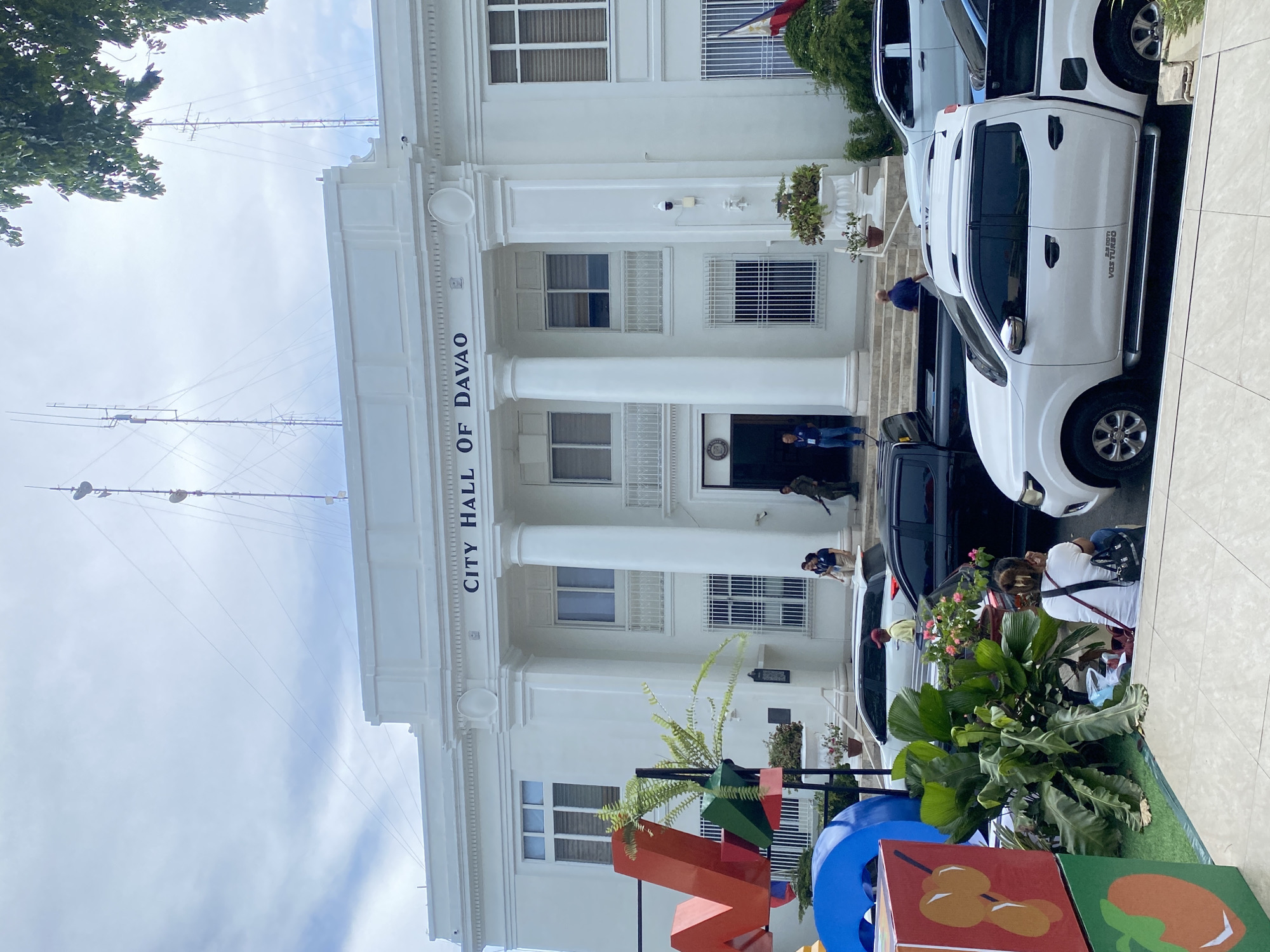 The image shows the City Hall of Davao, located in Davao City, Philippines. The building is a white, neoclassical structure with large columns and arched windows. In front of the building, several cars are parked, including white pickup trucks and a black SUV. Some people are visible on the steps of the city hall and near the vehicles. Colorful, large letter sculptures are present in the foreground. The sky is cloudy, and trees are visible on the right side of the image.