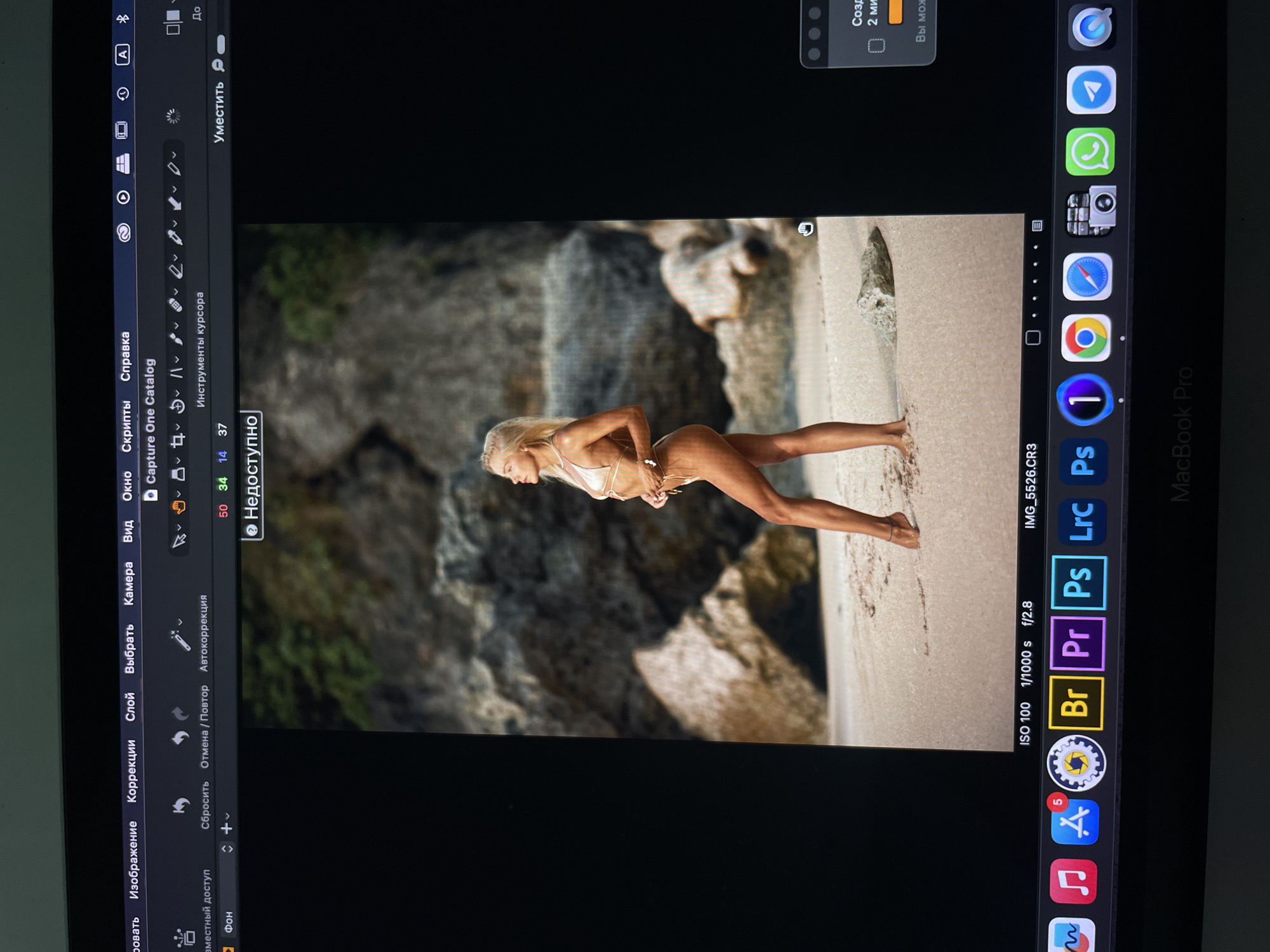 The image shows a laptop screen displaying photo editing software. The software, Capture One Catalog, is open, and an image of a woman in a bikini standing on a beach is visible in the editing interface. The ISO, shutter speed, and aperture are 100, 1/1000s, and f/2.8. The laptop's dock shows various applications, including a browser, Premiere Pro, and Photoshop. The top menu bar has standard options like "Image", "Correction", and "Window", displayed in Russian.
The location in the city of Kazan, Russia is not possible to determine from the image.