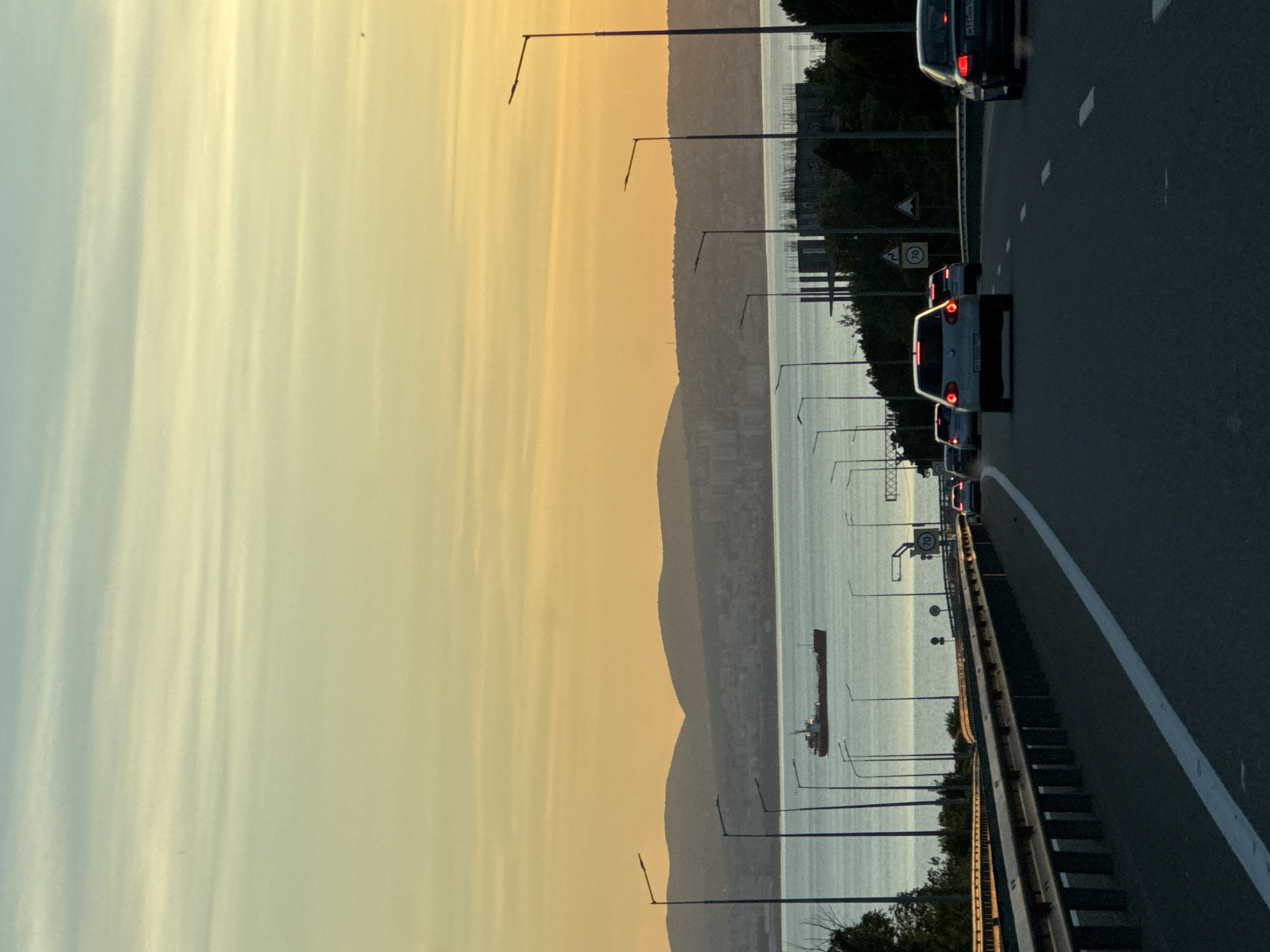 This detailed description outlines the visual elements present in the image, set in the city of Kabardinka, Russia.
The image captures an elevated view from a multi-lane highway at either sunrise or sunset, looking across a large body of water towards distant land. The sky exhibits a prominent gradient, transitioning from a pale blue-green at the top to a warm yellow and orange near the horizon, streaked with delicate horizontal cirrus cloud formations.
On the distant horizon, a series of dark, undulating mountain ranges are silhouetted against the bright sky. Below these mountains, across the water, an urbanized area is discernible as a lighter grey band, featuring numerous buildings.
The mid-ground is dominated by a calm, blue-grey body of water, likely a sea or bay, consistent with the Black Sea coastline near Kabardinka. A dark-hulled vessel, appearing to be a tanker or cargo ship, is positioned horizontally on the water slightly left of center, seemingly stationary.
In the foreground and mid-ground, the asphalt highway curves gently to the right. A prominent dark metal guardrail with red reflectors lines the left side of the road. Numerous tall, dark streetlights with single arms are silhouetted against the sky and water, extending over the road and along its edges. These lights are unilluminated.
Traffic signs are visible on the right side of the road: a circular yellow sign with "70" in black, indicating a speed limit of 70 km/h; a triangular white warning sign with a red border and an exclamation mark; and another triangular white warning sign with a red border depicting a winding road. Dark green foliage borders the road, particularly on the right. Further to the right, a construction site is evident with visible concrete structural elements (columns and walls) silhouetted against the water.
Several cars are traveling away from the viewer on the highway. A light-colored (likely silver or grey) Volkswagen Golf is in the middle lane, displaying illuminated red brake lights. Its license plate reads `H112BO 152`. To its right, in the adjacent lane, a dark-colored sedan (possibly an Audi A4) also has illuminated red brake lights, with a license plate reading `M117HH 152`. Additional vehicles appear as dark shapes with red brake lights further down the road. No individuals are visible in the scene.