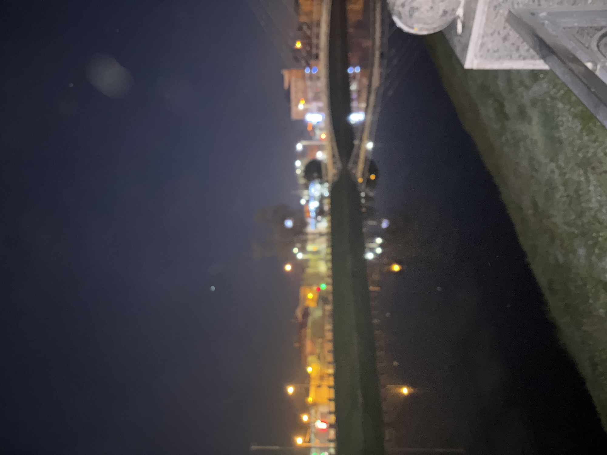 The image shows a night view in Kharkiv, Ukraine. The lights of a bridge and city are reflected in water, possibly a river or canal. In the foreground, there is a stone or concrete structure with some vegetation. The night sky is visible.